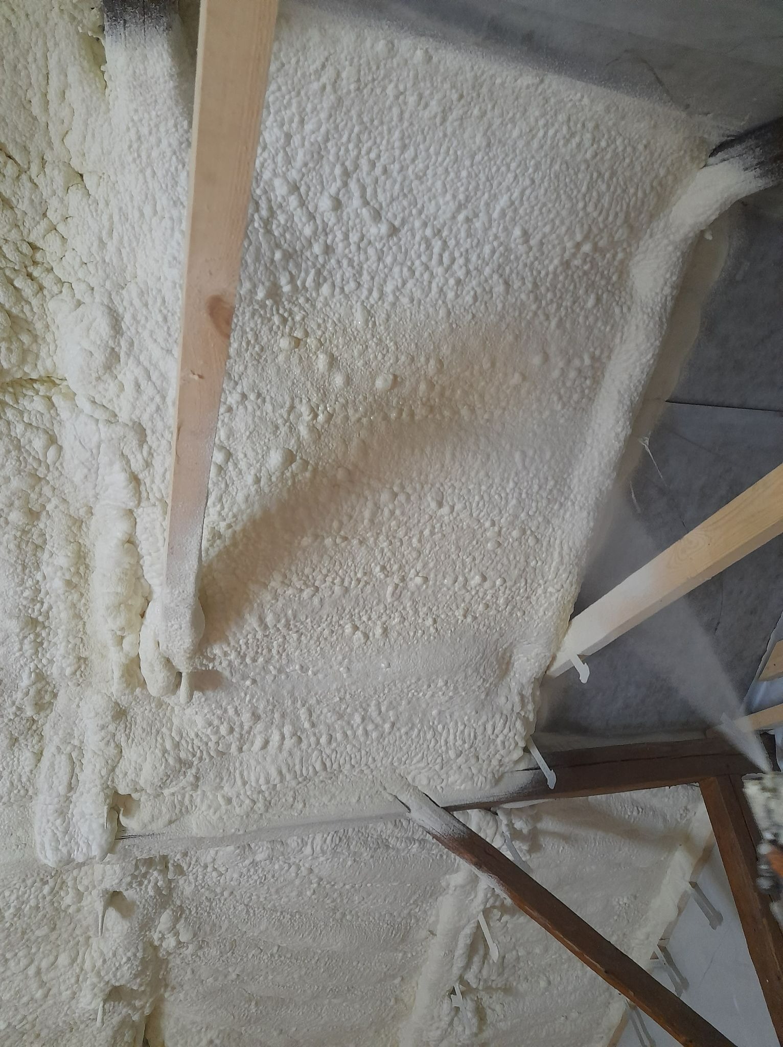 Świeżo nałożona, jasna warstwa izolacyjna na poddaszu, widoczne drewniane elementy konstrukcyjne i fragment dyszy aplikatora.