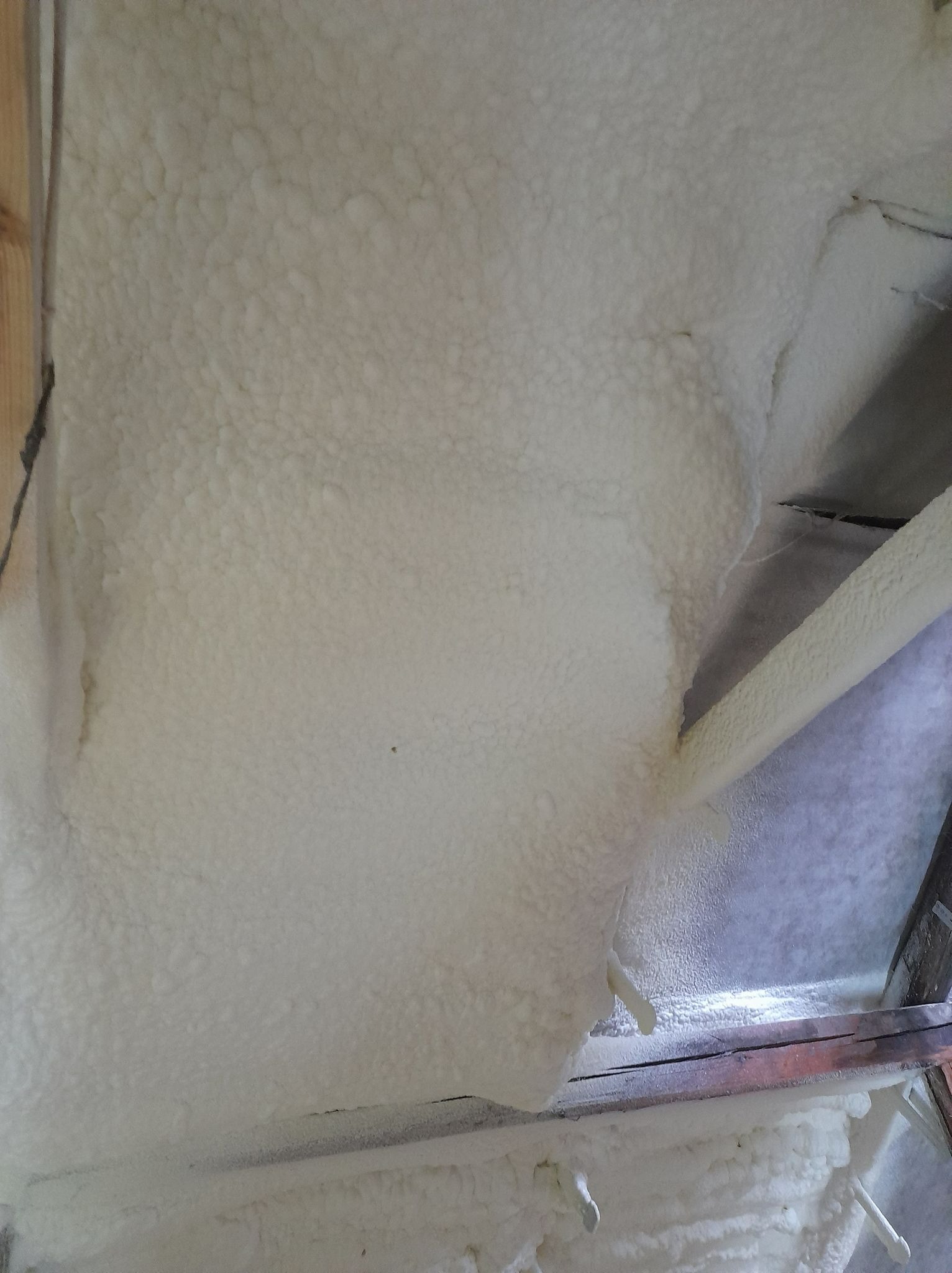 Świeżo nałożona warstwa izolacyjna z pianki na poddaszu, widoczne drewniane elementy konstrukcyjne i nierównomierna powierzchnia piany.