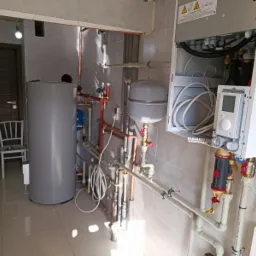 Wnętrze kotłowni z zamontowanym zasobnikiem ciepłej wody, pompą ciepła z widocznym sterownikiem i licznymi rurami miedzianymi oraz plastikowymi.