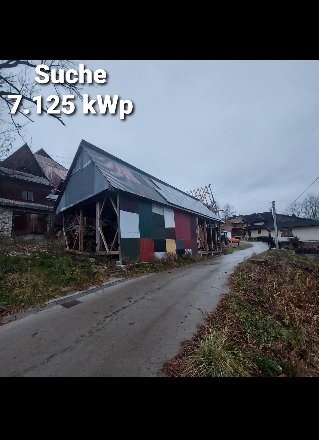 Stary budynek gospodarczy z częściowo zamontowanymi panelami słonecznymi na dachu i kolorowymi płytami na ścianach, widok z poziomu drogi w pochmurny dzień.