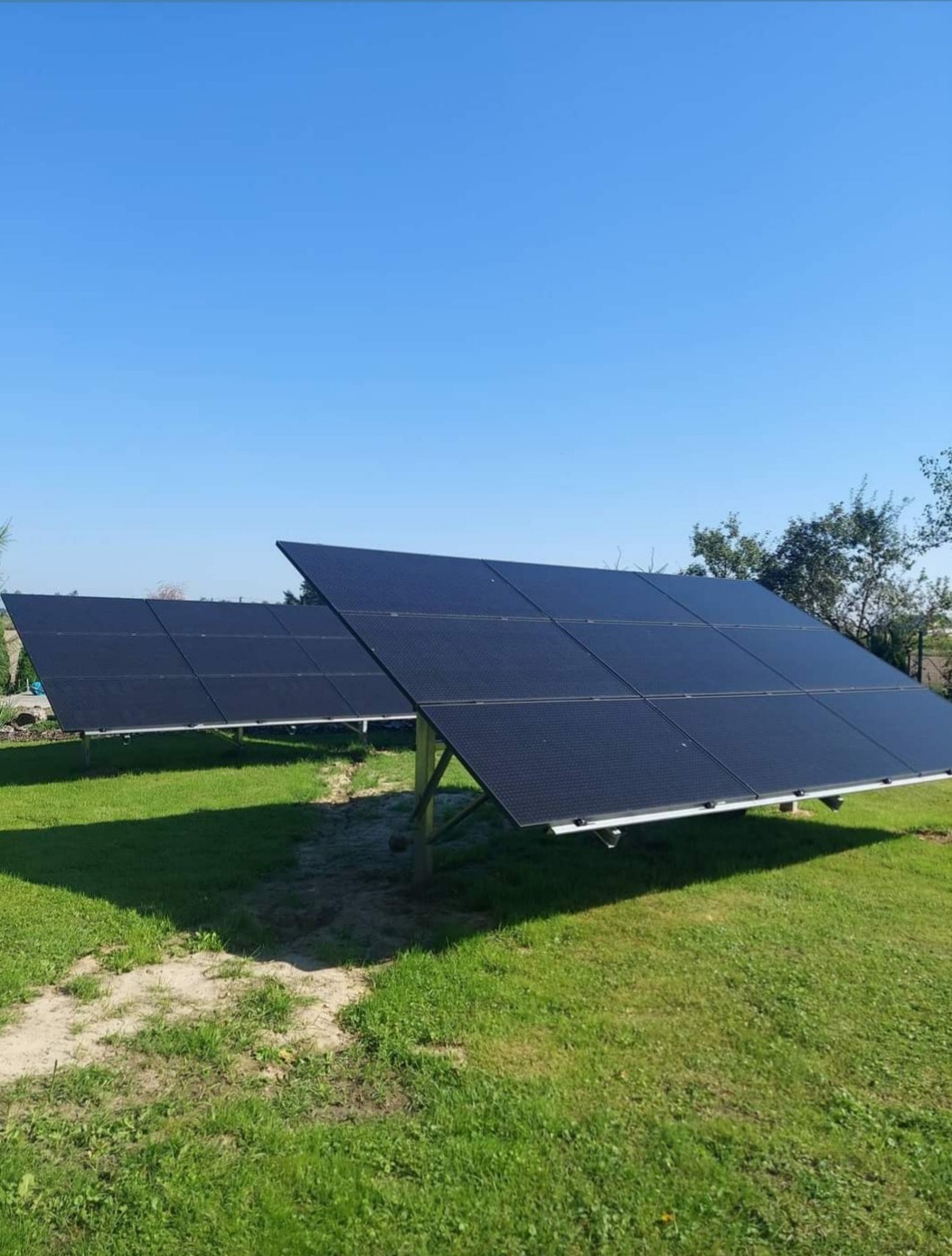 Dwa panele słoneczne ustawione na trawniku pod błękitnym niebem, widoczne ślady po montażu w postaci uschniętej trawy wokół podstawy konstrukcji.