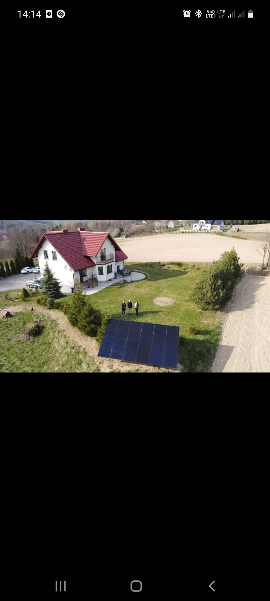 Widok z góry na panele fotowoltaiczne ustawione na trawniku przed domem z czerwoną dachówką, obok których stoją trzy osoby.