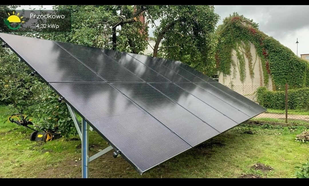 Czarny panel fotowoltaiczny na metalowej konstrukcji ustawiony na trawniku, w tle budynek porośnięty bluszczem i kosiarka do trawy.