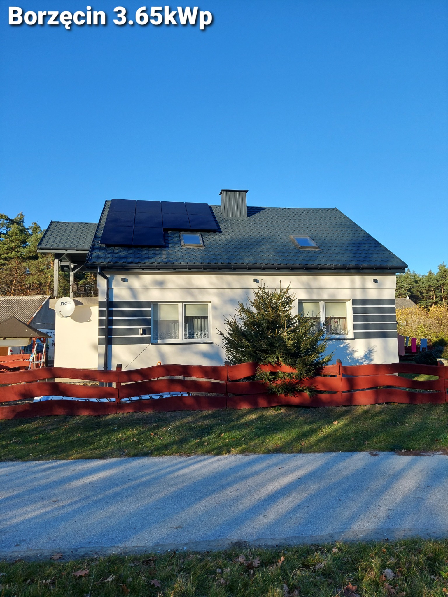 Dom jednorodzinny z panelami słonecznymi na dachu, widok z ulicy w słoneczny dzień, czerwone ogrodzenie i świerk przed budynkiem.