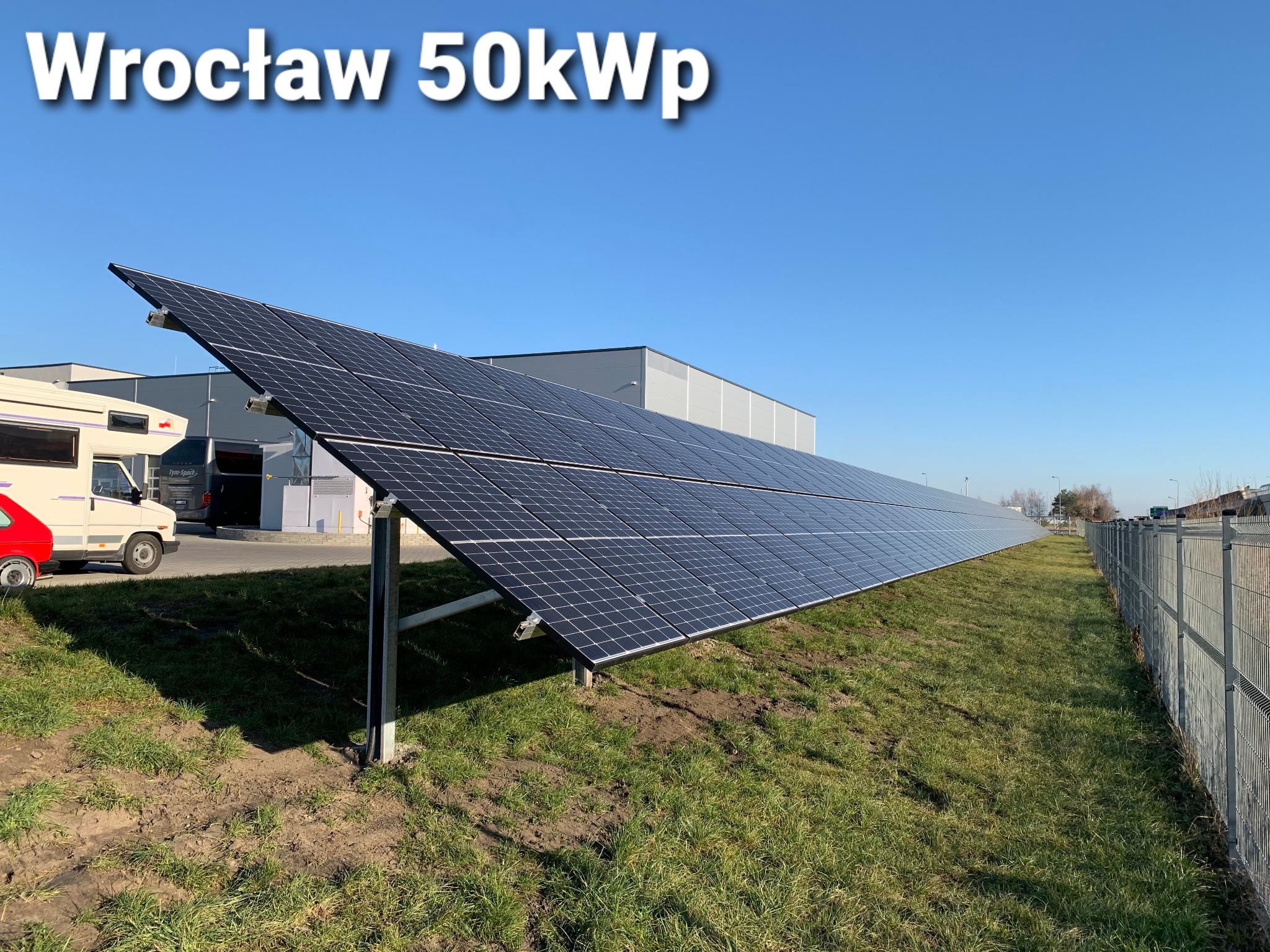 Instalacja paneli słonecznych o mocy 50kW we Wrocławiu, widoczna z perspektywy gruntu, z budynkiem przemysłowym i pojazdami w tle.