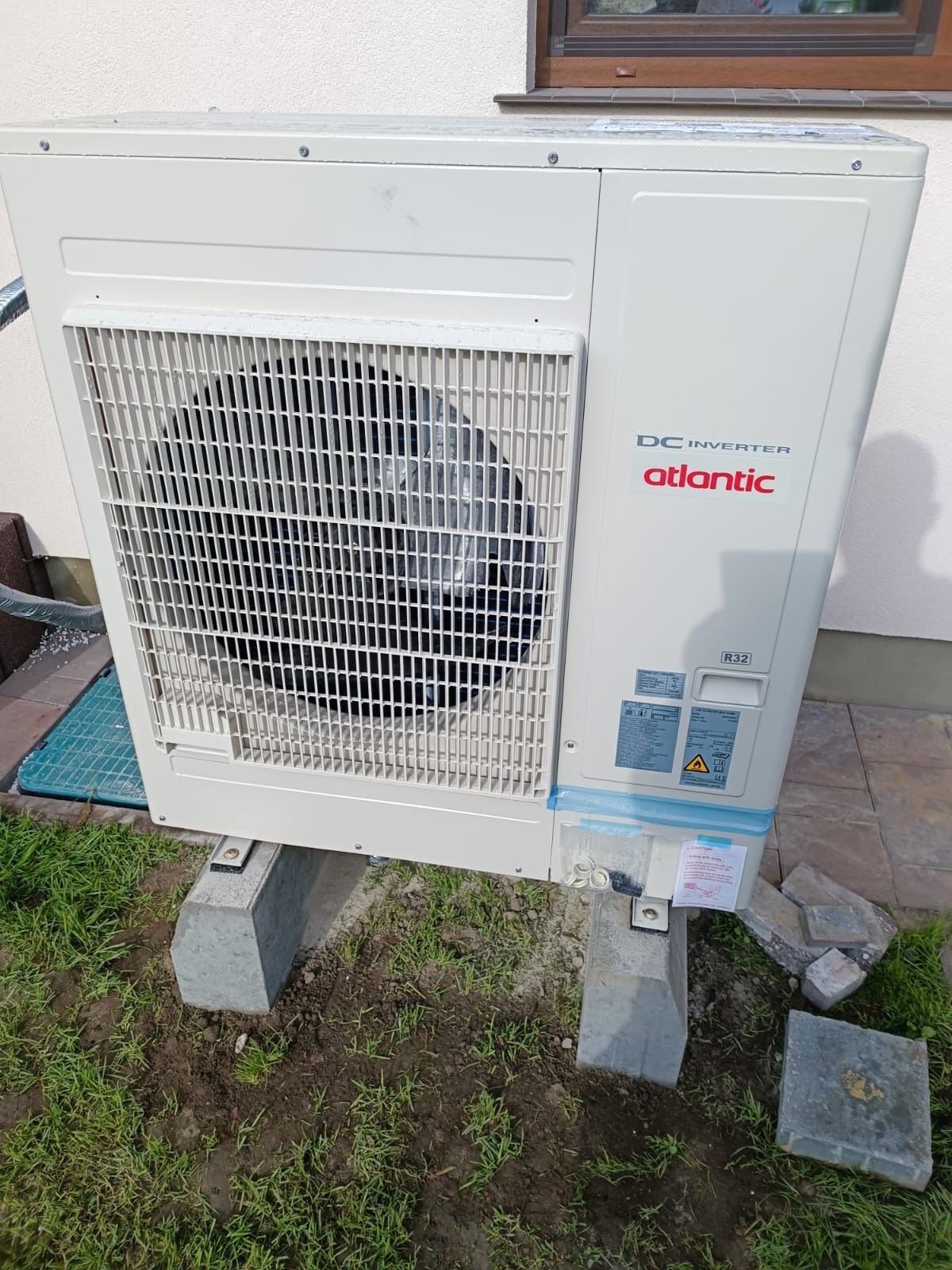 Zewnętrzna jednostka pompy ciepła Atlantic DC Inverter, zamontowana na betonowych wspornikach na trawniku, z widocznymi naklejkami informacyjnymi i elementami montażowymi.