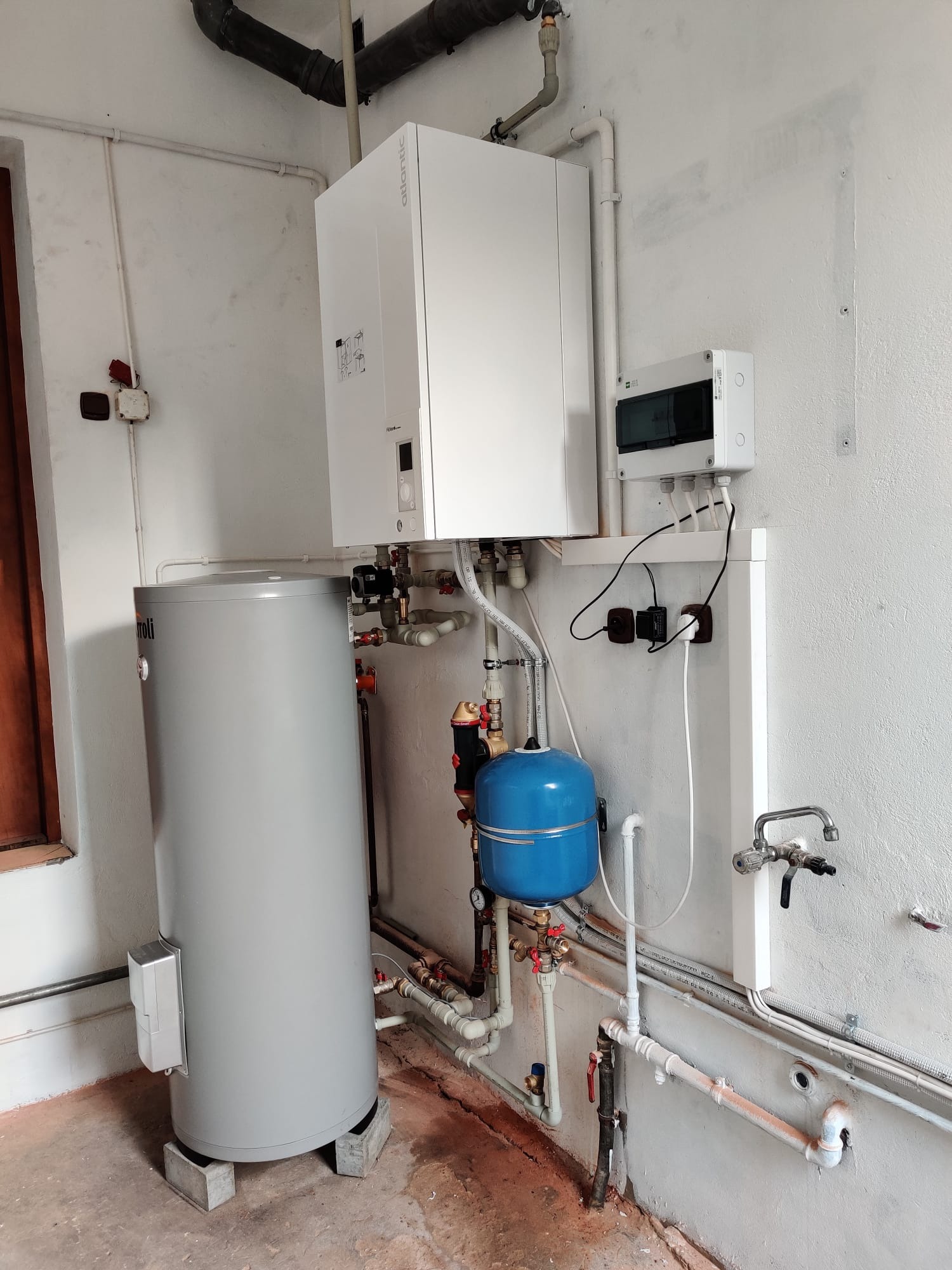 Biały piec grzewczy Atlantic zamontowany nad szarym zasobnikiem wody, z niebieskim naczyniem przeponowym, instalacją hydrauliczną i elektryczną na tle białej ściany w pomieszczeniu technicznym.