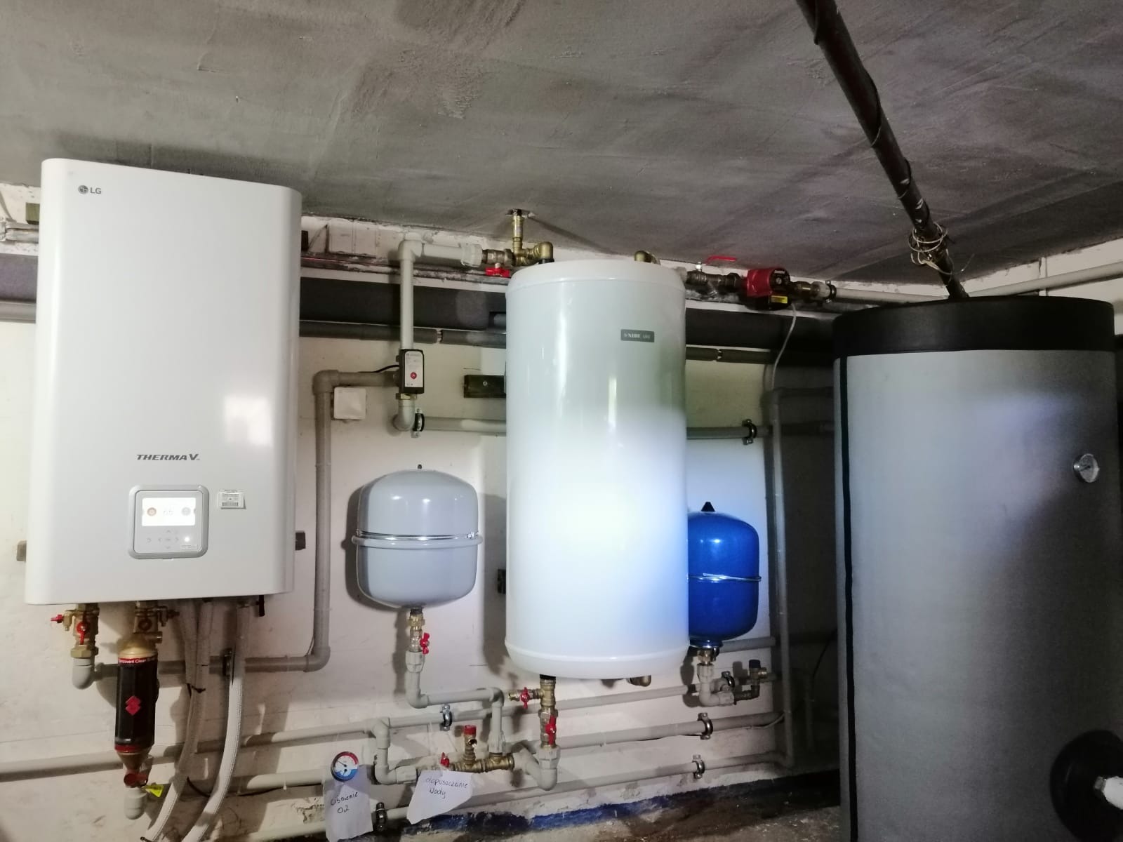 Instalacja pompy ciepła LG Therma V z zasobnikami wody o różnej pojemności i kolorach, widoczne rury i zawory, w pomieszczeniu technicznym z surowym betonowym sufitem.