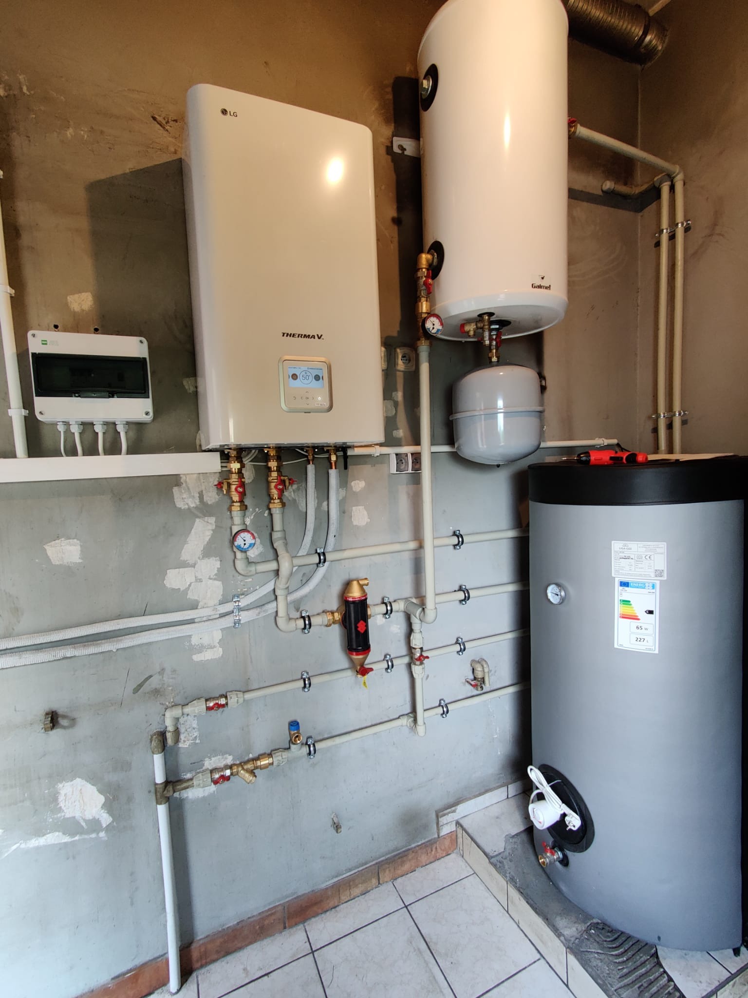 Instalacja grzewcza w pomieszczeniu technicznym: wiszący biały moduł LG Therma V, biały zasobnik Galmet, szary zbiornik akumulacyjny, rury i zawory, widoczne narzędzia na zbiorniku, surowe ściany...