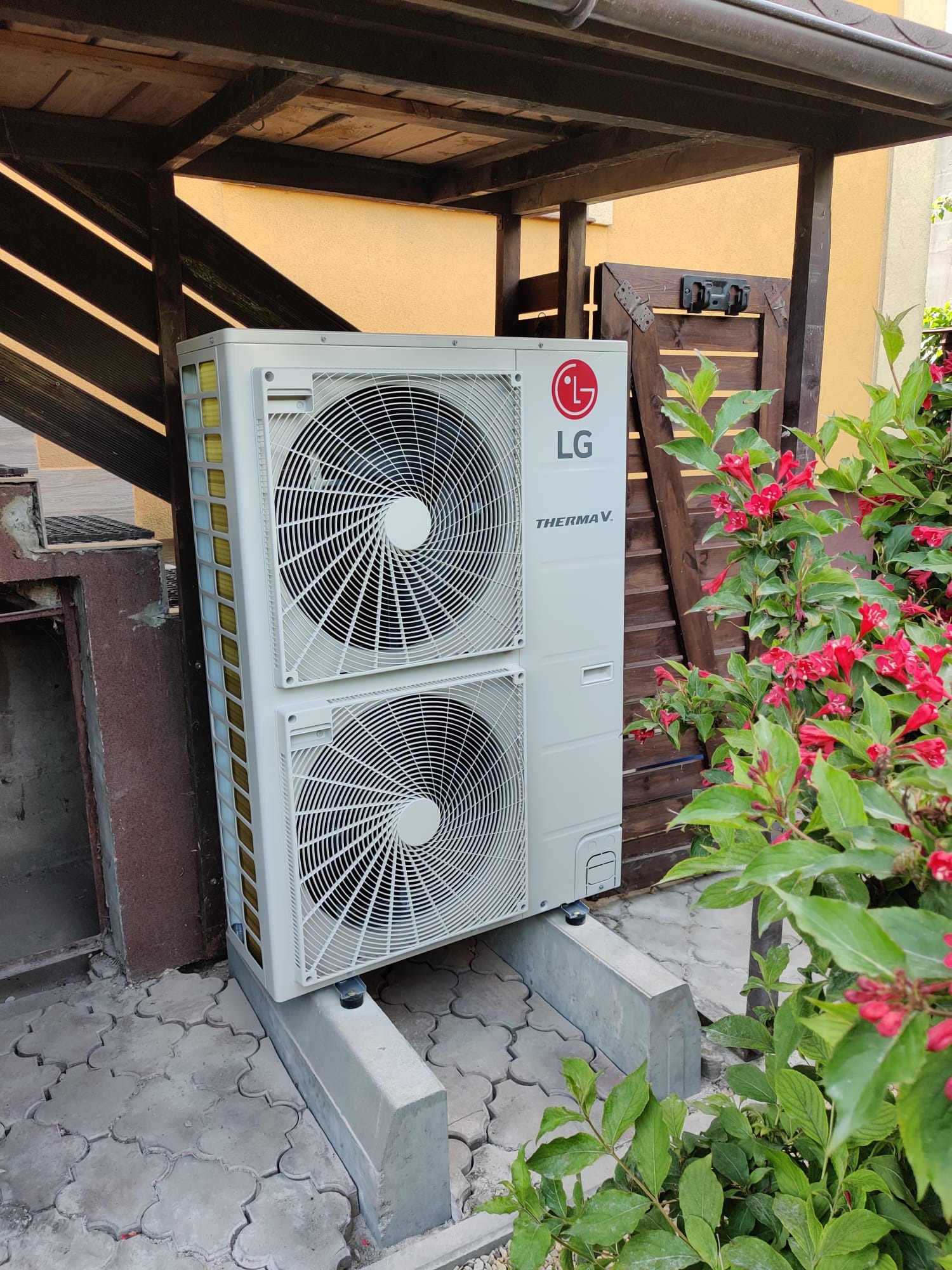 Biała, dwuwentylatorowa pompa ciepła LG Therma V umieszczona na betonowych wspornikach, obok kwitnącego krzewu z czerwonymi kwiatami, na tle drewnianej konstrukcji i ściany budynku.