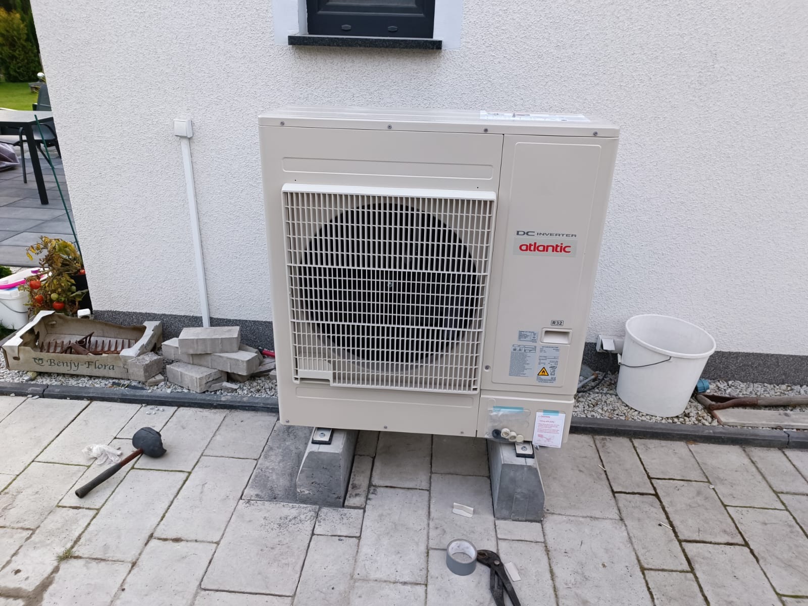 Zewnętrzna jednostka pompy ciepła Atlantic DC Inverter na betonowych podstawach, widoczne narzędzia: młotek, taśma i szczypce, obok wiadro i skrzynka z roślinami.
