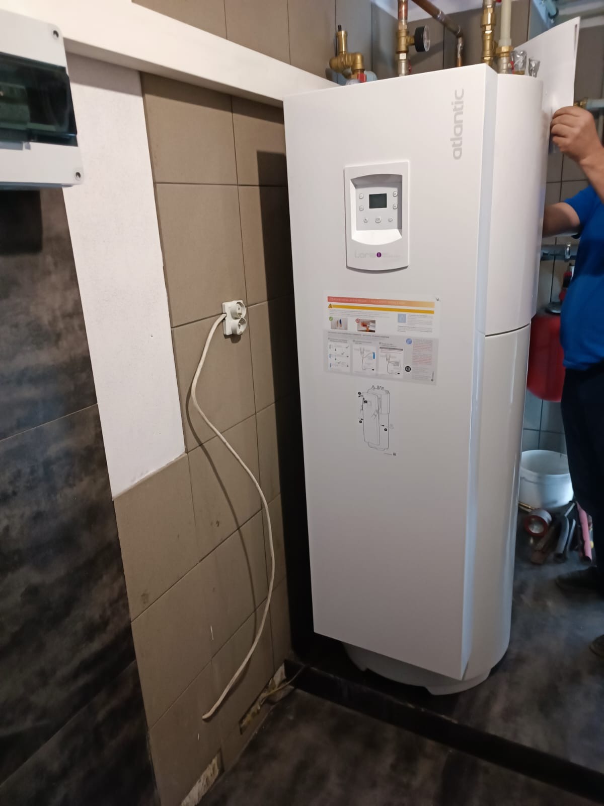 Biała pompa ciepła Atlantic z panelem sterowania, zamontowana w pomieszczeniu z beżowymi płytkami, widoczne rury instalacji i fragment osoby w niebieskiej koszulce.