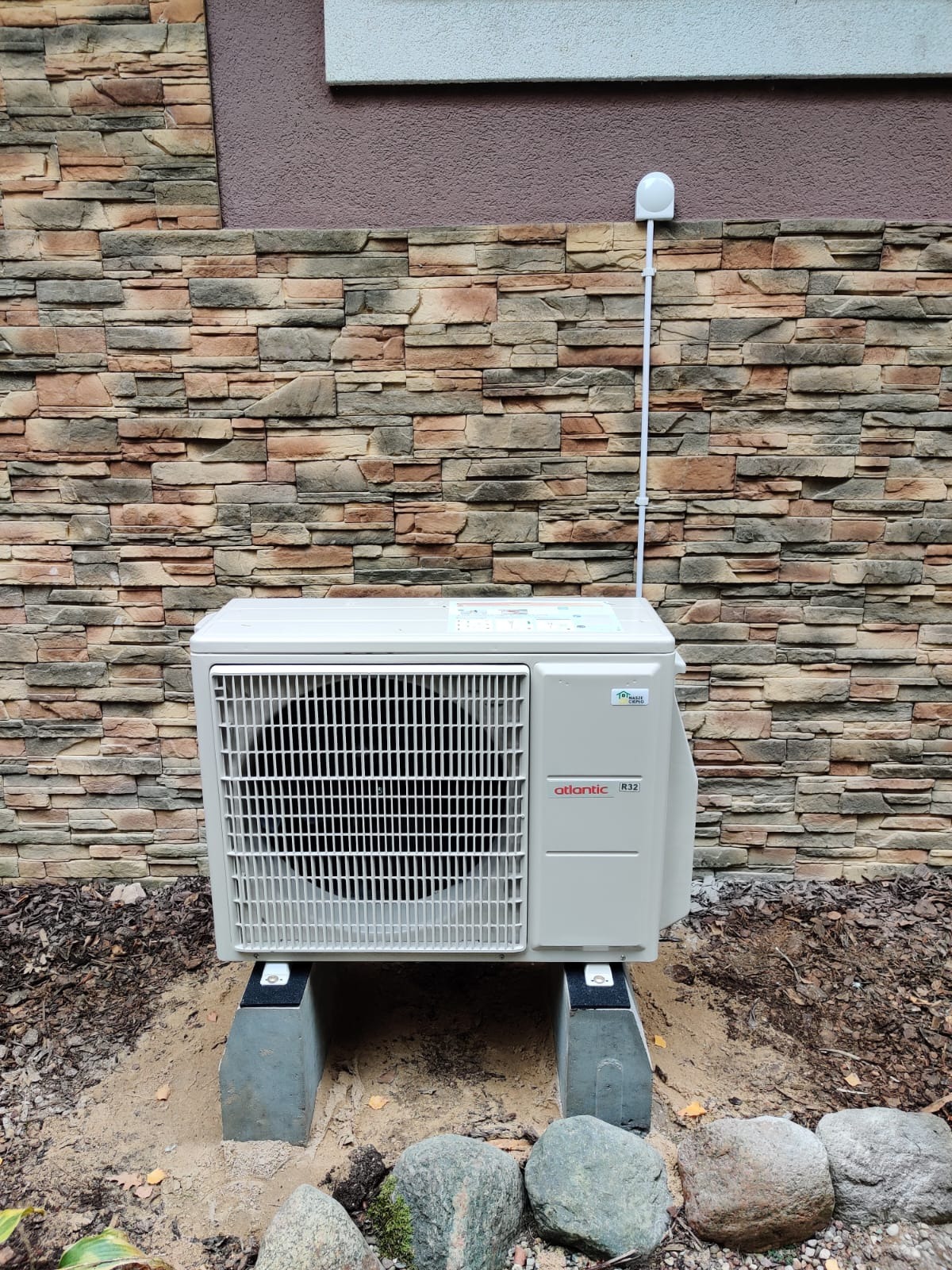 Kompaktowa jednostka zewnętrzna klimatyzacji marki Atlantic R32, umieszczona na betonowych wspornikach przed elewacją z kamienia dekoracyjnego, z widoczną instalacją elektryczną.