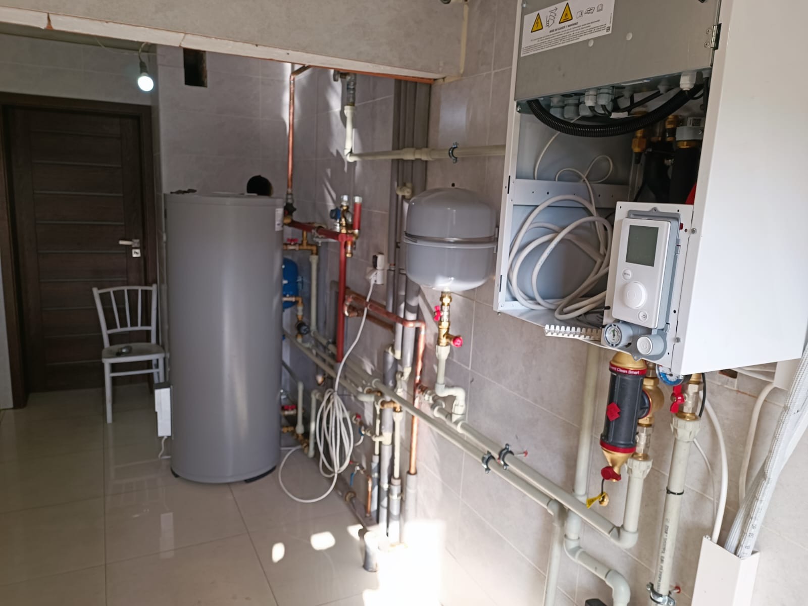 Wnętrze kotłowni z zamontowanym zasobnikiem ciepłej wody, pompą ciepła z widocznym sterownikiem i licznymi rurami miedzianymi oraz plastikowymi.