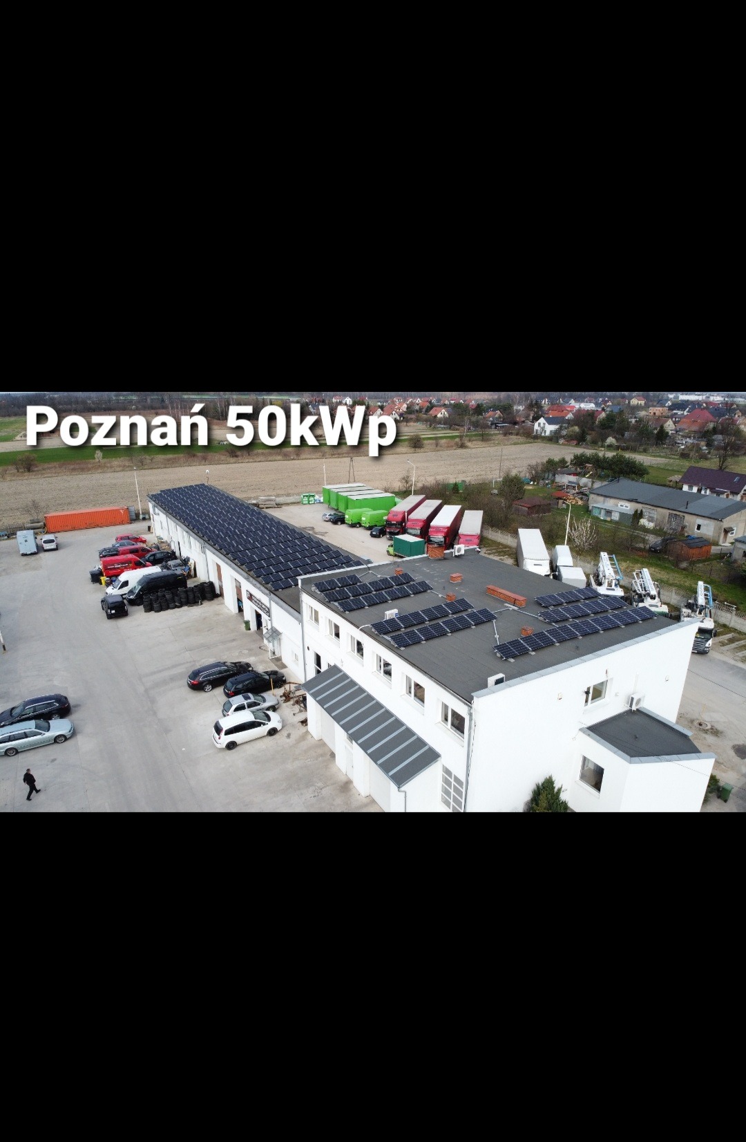 Widok z lotu ptaka na biały budynek z panelami słonecznymi na dachu, z napisem 'Poznań 50kWp' nałożonym na obraz, zaparkowane samochody i ciężarówki w pobliżu.