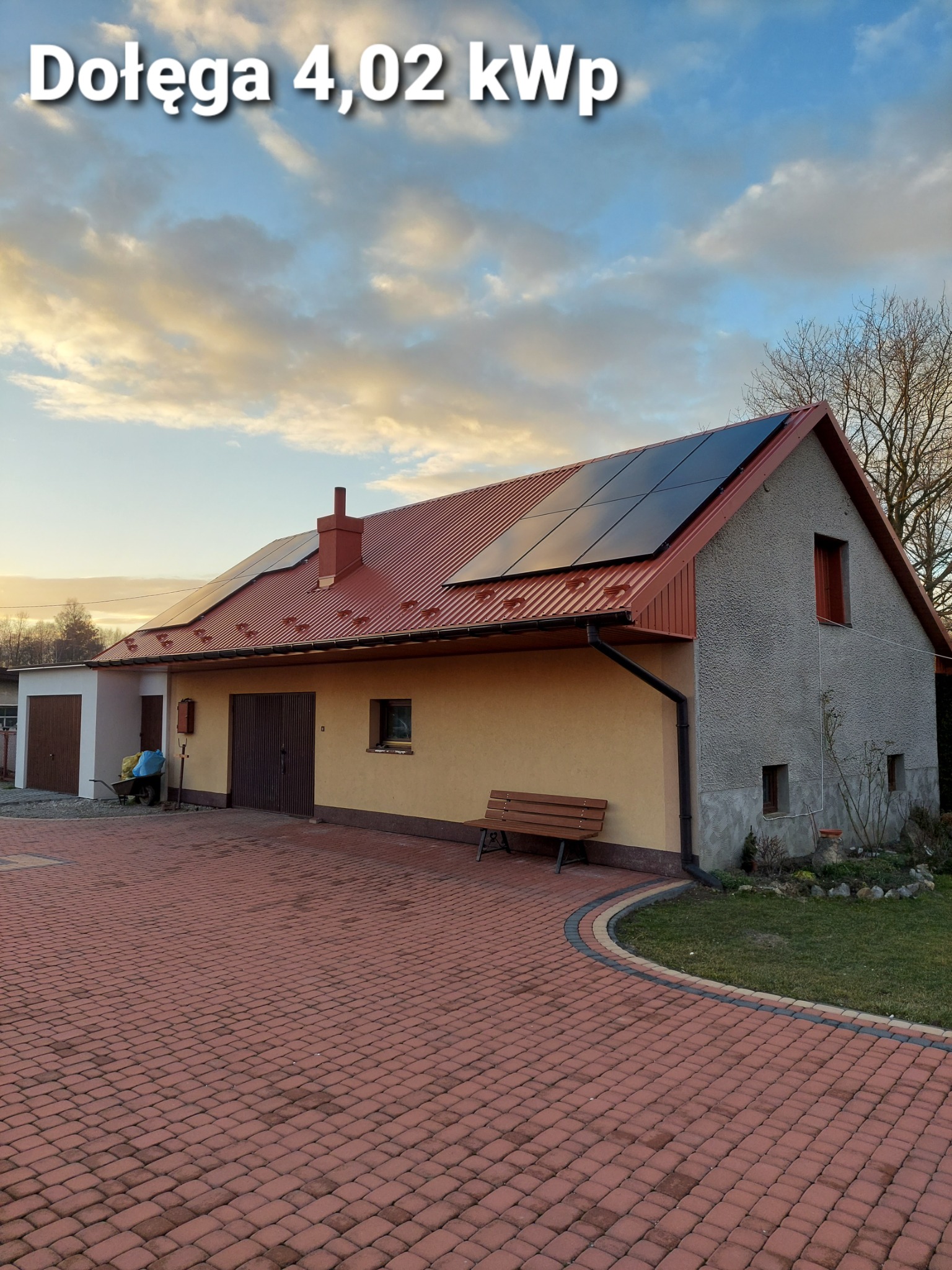 Dom jednorodzinny z panelami słonecznymi na czerwonym dachu, widok z brukowanego podjazdu o zmierzchu, z napisem 'Dołęga 4,02 kWp' w górnej części zdjęcia.