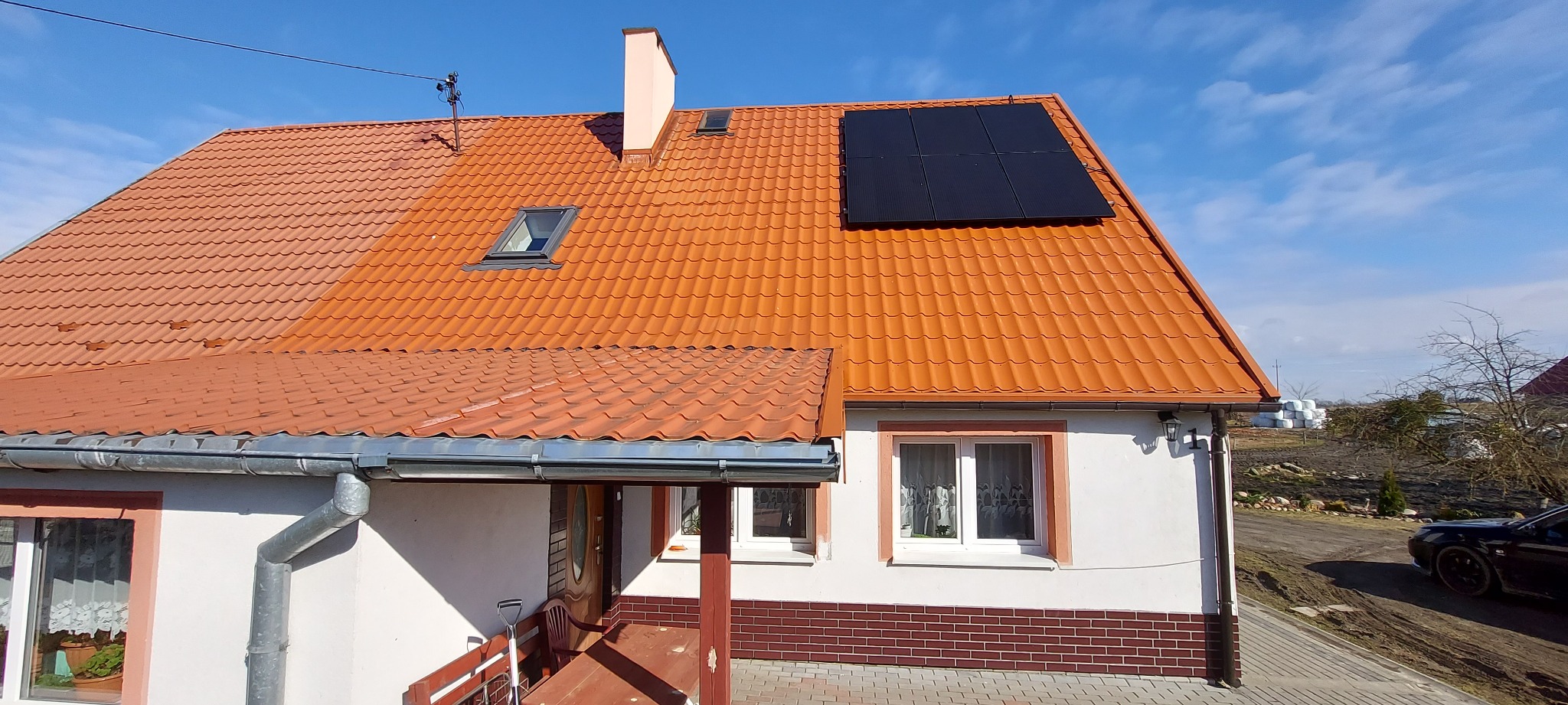 Dom jednorodzinny z pomarańczową dachówką i czarnymi panelami słonecznymi na dachu, widok z poziomu gruntu, błękitne niebo w tle.