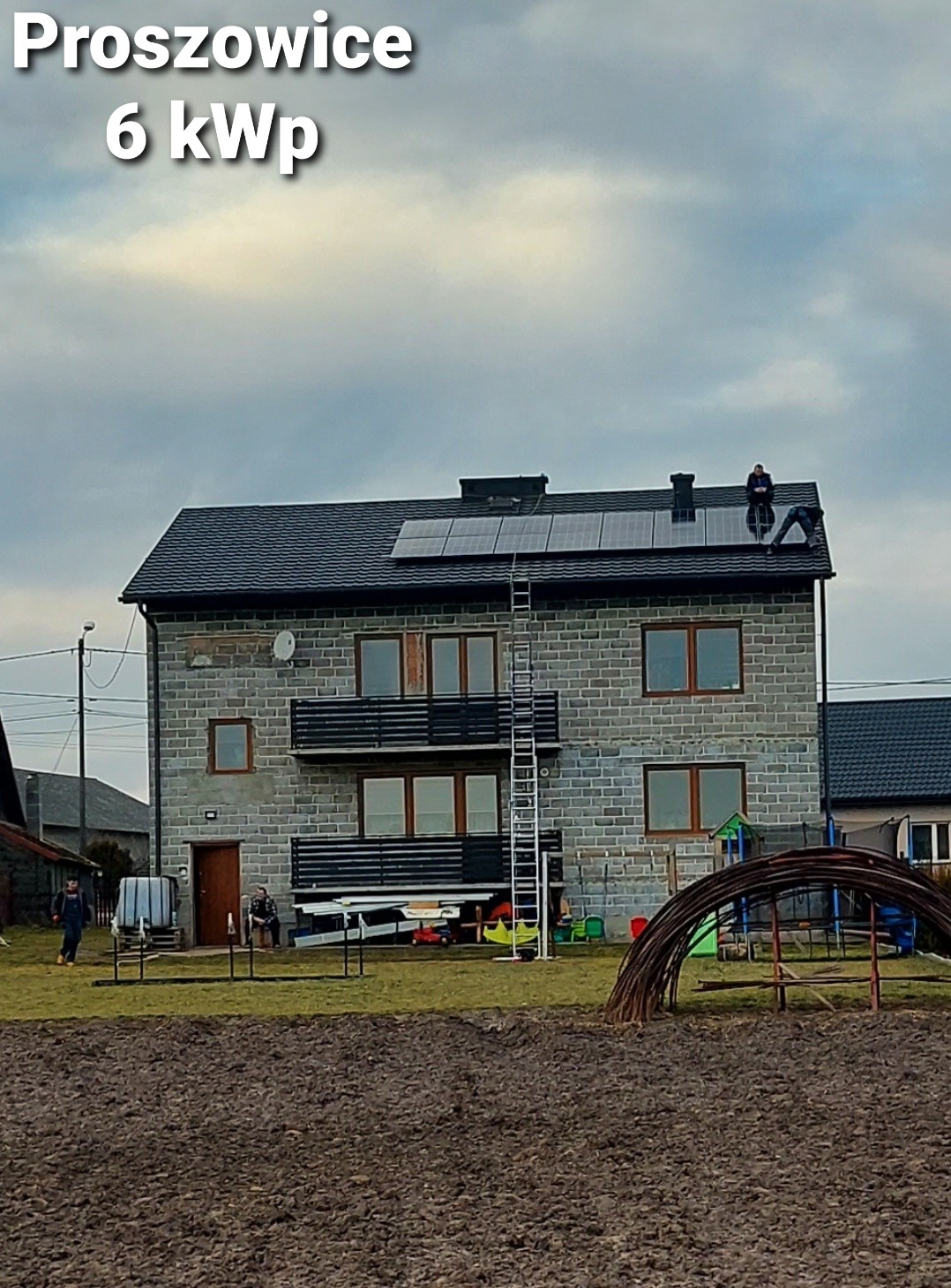 Montaż paneli słonecznych na dachu dwupiętrowego domu z szarej cegły w Prosowicach, widoczna drabina i dwaj monterzy na dachu, w tle pochmurne niebo i ogród z elementami placu zabaw.
