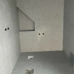 Wnętrze łazienki w trakcie wykańczania, ściany i podłoga pokryte szarymi płytkami z drobnym wzorem, widoczne otwory na gniazdka elektryczne i odpływ pod prysznic.