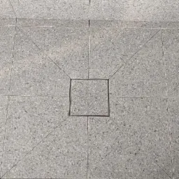 Szara podłoga z płytek z odpływem liniowym w kształcie kwadratu, widoczne stopy w zielonych klapkach.