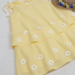 Pracownia Krawiecka CASSIE Jolanta Szymczak - Żółta, dwuwarstwowa sukienka dziecięca ozdobiona białymi stokrotkami, leżąca obok słomkowego kapelusza z wiankiem z polnych kwiatów.