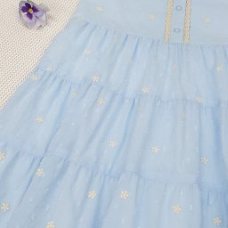 Pracownia Krawiecka CASSIE Jolanta Szymczak - Błękitna, warstwowa sukienka dziewczęca z kołnierzykiem i koronkowym wykończeniem, ozdobiona haftowanymi kwiatkami, leżąca na białej tkaninie w towarzystwie hortensji i bratków.