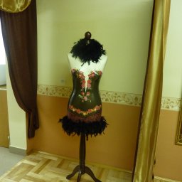 Pracownia Krawiecka CASSIE Jolanta Szymczak - Manekin krawiecki z dekoracyjnym gorsetem w kwiaty i pióra, stojący w pomieszczeniu z beżowymi ścianami, drewnianą podłogą i złotą zasłoną