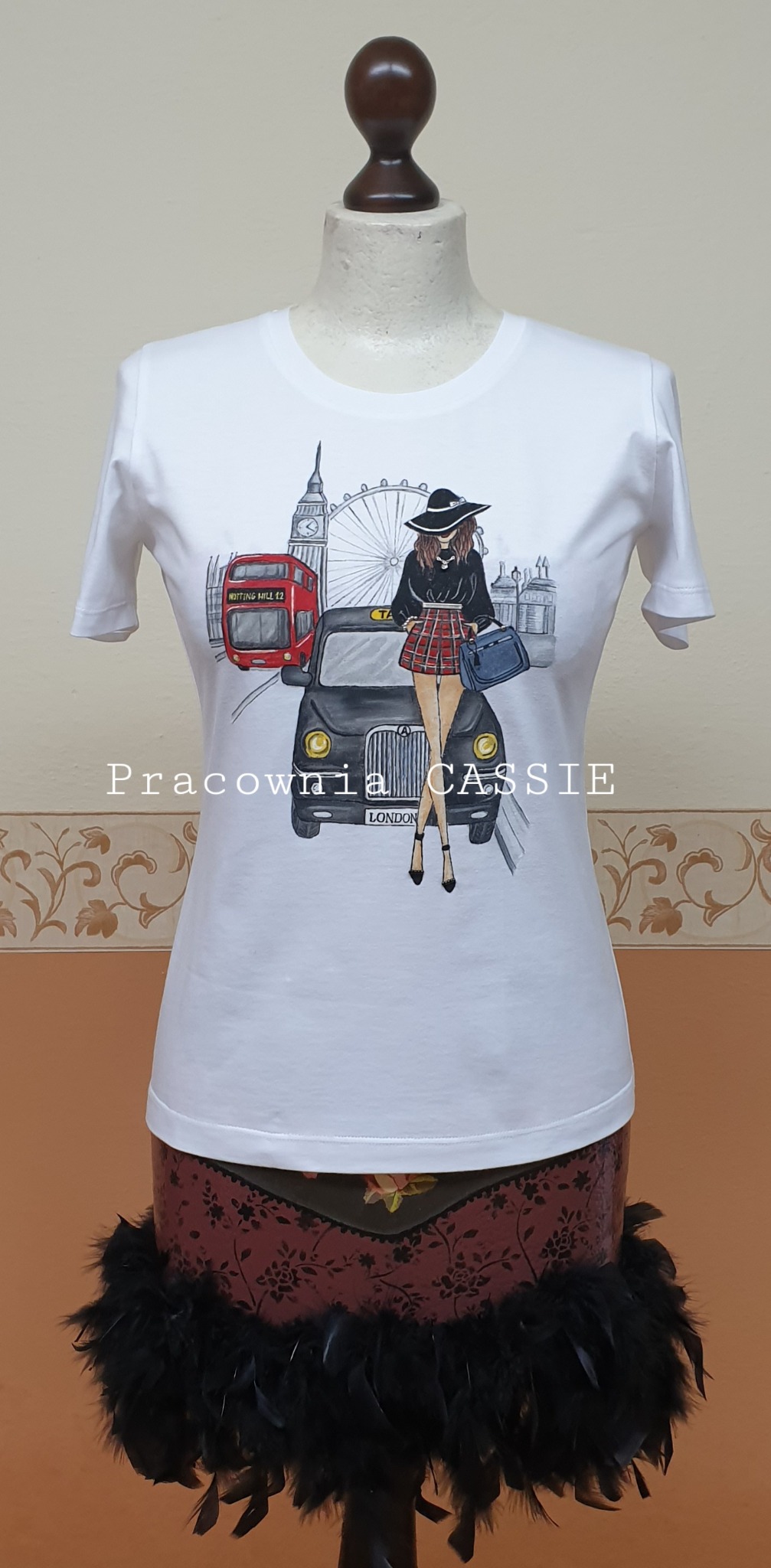 Biały t-shirt z ręcznie malowaną ilustracją londyńskiej taksówki, czerwonego autobusu, Big Bena i kobiety w kapeluszu, prezentowany na manekinie.