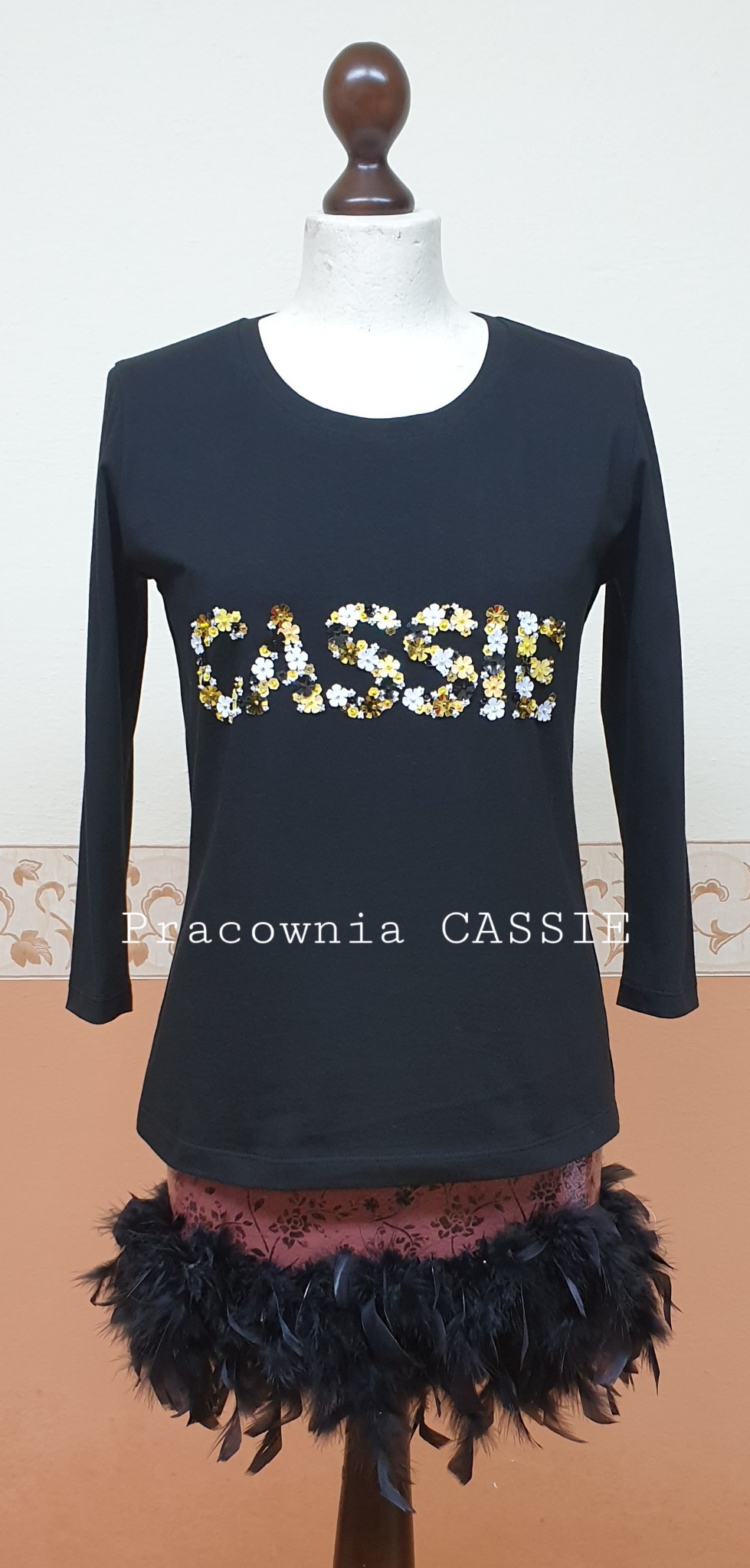 Czarna bluzka z długim rękawem, z napisem 'CASSIE' wykonanym z cekinów w kolorach białym, złotym i czarnym, prezentowana na manekinie z dekoracją z czarnych piór u dołu.