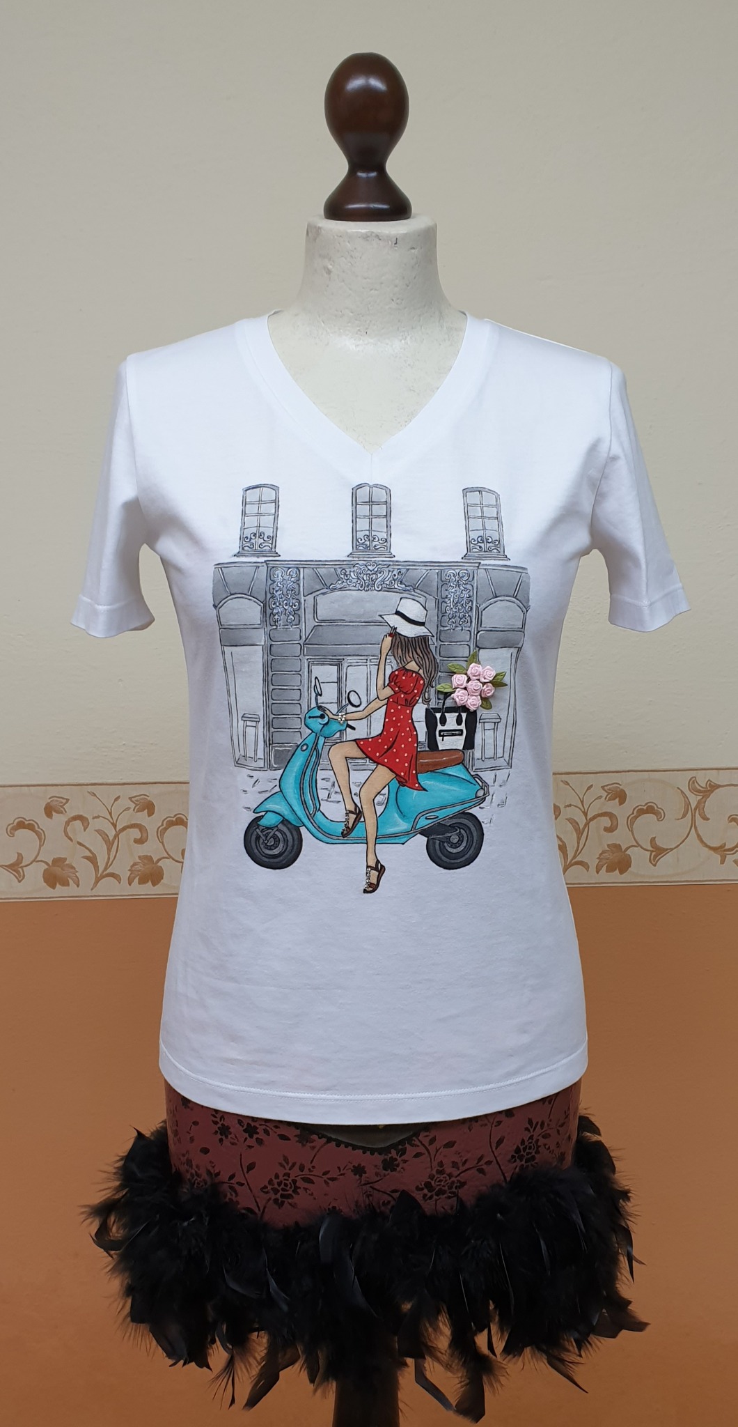 Biała koszulka z nadrukiem przedstawiającym kobietę na turkusowym skuterze przed budynkiem, prezentowana na manekinie w Firleju.