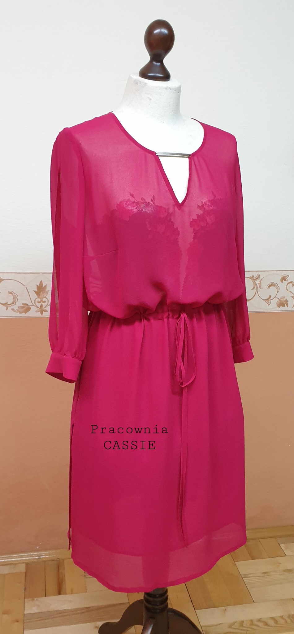 Manekin prezentuje zwiewną, malinową sukienkę z długimi, transparentnymi rękawami, ze srebrnym detalem przy dekolcie w kształcie litery V i wiązaniem w pasie. Na dole sukienki widoczny napis...