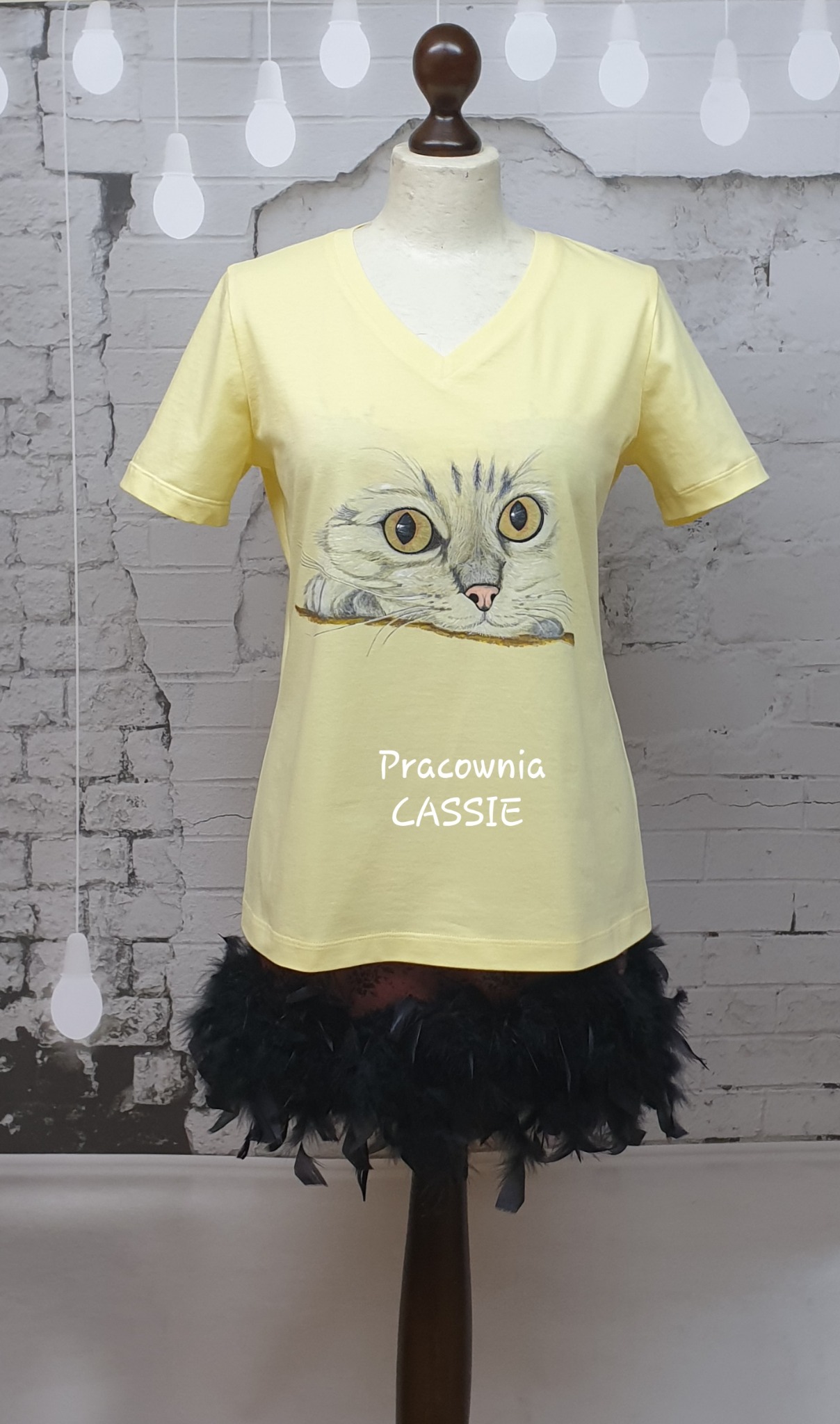 Żółta koszulka z nadrukiem kota na manekinie, z dopiętą spódnicą z piór, na tle ceglanej ściany z dekoracyjnymi żarówkami.