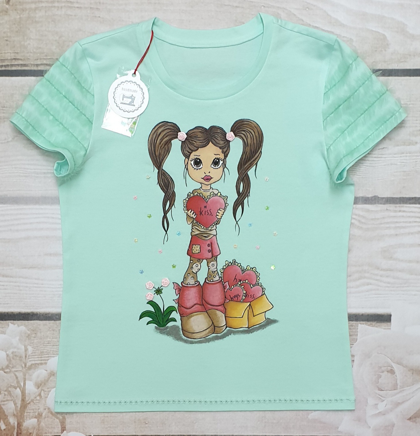 Jasnomiętowa koszulka dziecięca z ręcznie malowaną dziewczynką trzymającą serce z napisem 'Kiss', obok pudełko z sercami, na drewnianym tle.