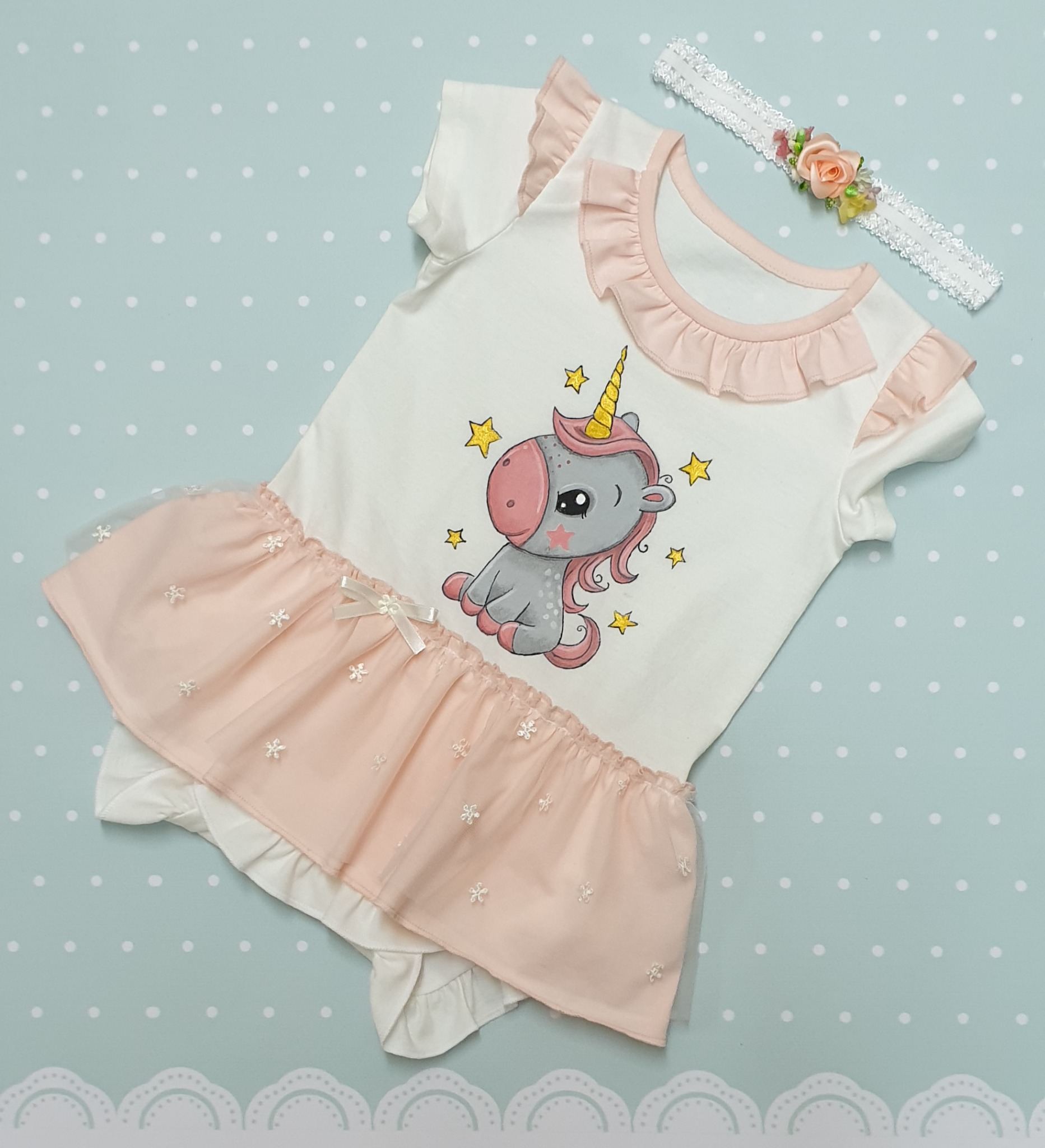 Białe body niemowlęce z krótkim rękawem i falbankami w kolorze pudrowego różu, zdobione rysunkiem szarego jednorożca i tiulową spódniczką, leżące obok opaski z kwiatkiem na tle w pastelowe kropki.
