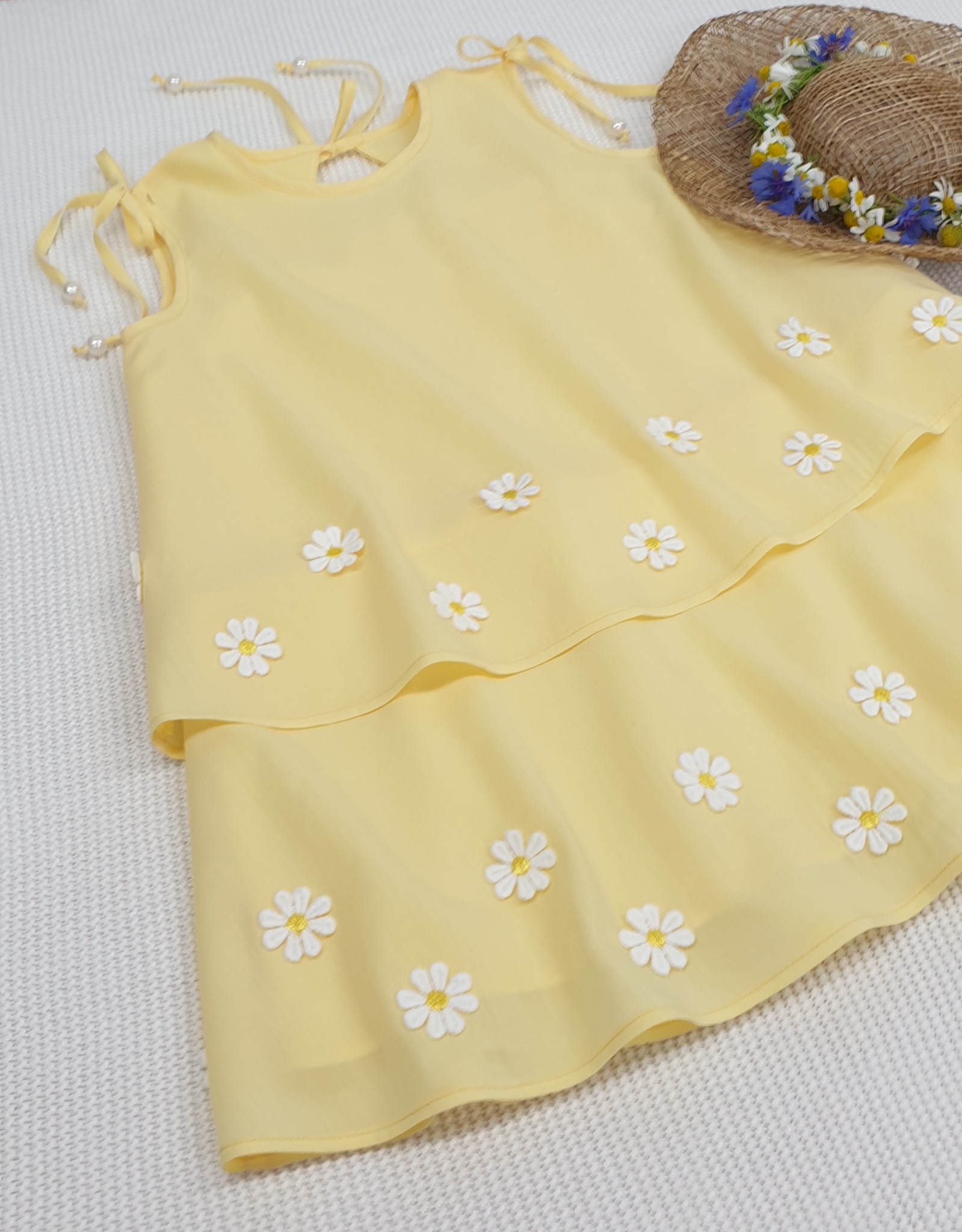 Żółta, dwuwarstwowa sukienka dziecięca ozdobiona białymi stokrotkami, leżąca obok słomkowego kapelusza z wiankiem z polnych kwiatów.