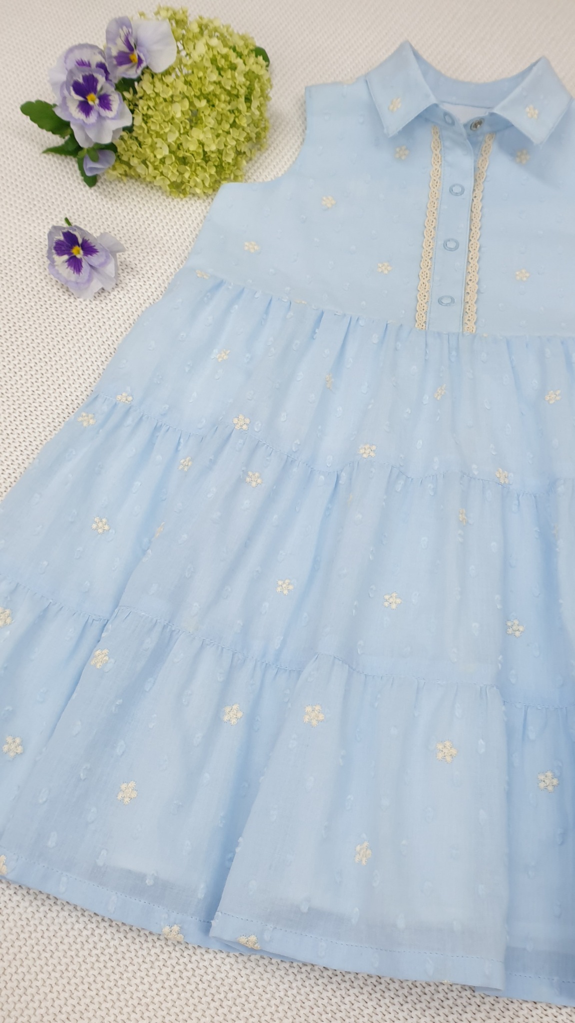 Błękitna, warstwowa sukienka dziewczęca z kołnierzykiem i koronkowym wykończeniem, ozdobiona haftowanymi kwiatkami, leżąca na białej tkaninie w towarzystwie hortensji i bratków.