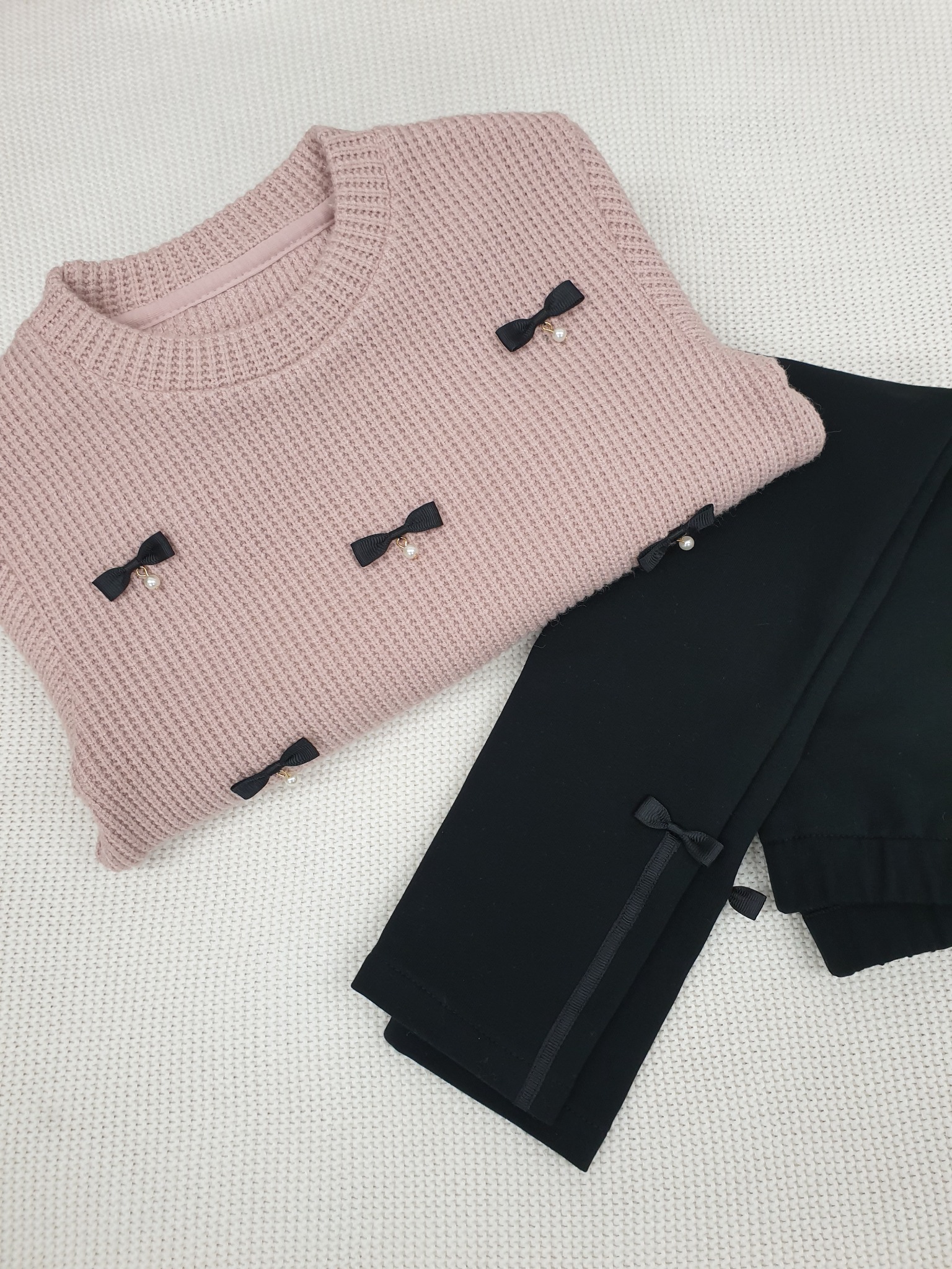Różowy sweter z dzianiny z czarnymi kokardkami i perłami, leżący obok czarnych spodni z kokardkami na dole.