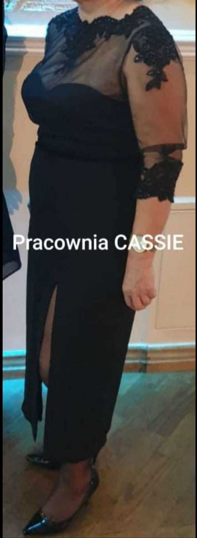 Czarna, elegancka suknia wieczorowa z koronkowymi zdobieniami na ramionach i dekolcie, z rozcięciem na nodze, prezentowana na osobie.