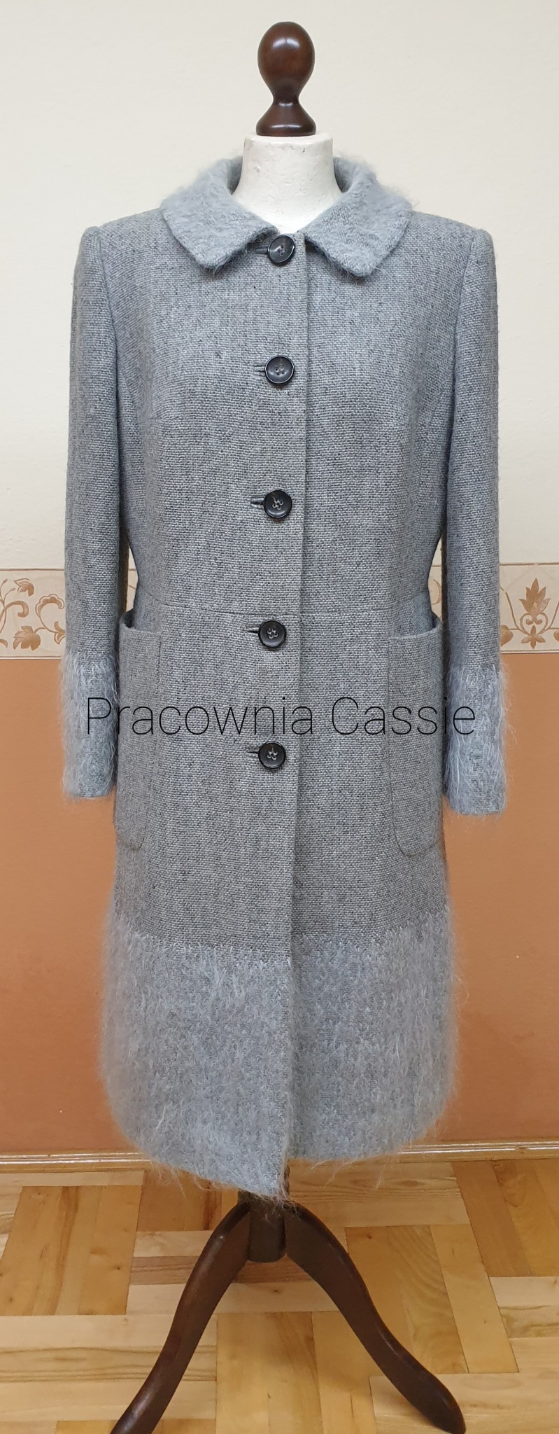 Szary płaszcz damski z wełnianymi wstawkami na manekinie, z trzema czarnymi guzikami i kieszeniami.