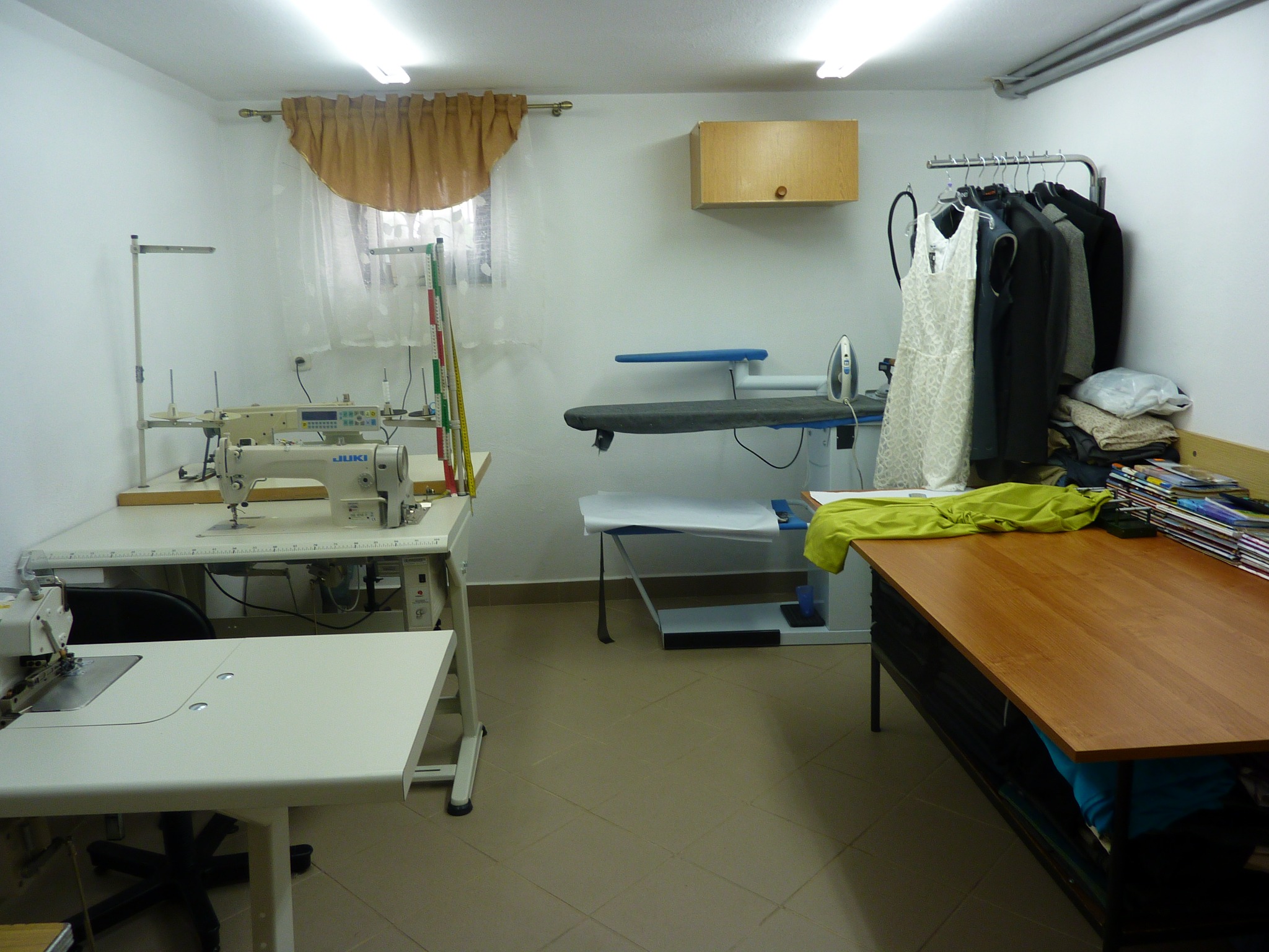 Wnętrze pracowni: dwie maszyny do szycia marki JUKI, deska do prasowania z żelazkiem, wieszak z ubraniami (sukienka koronkowa i marynarki) oraz stół z materiałami i magazynami.
