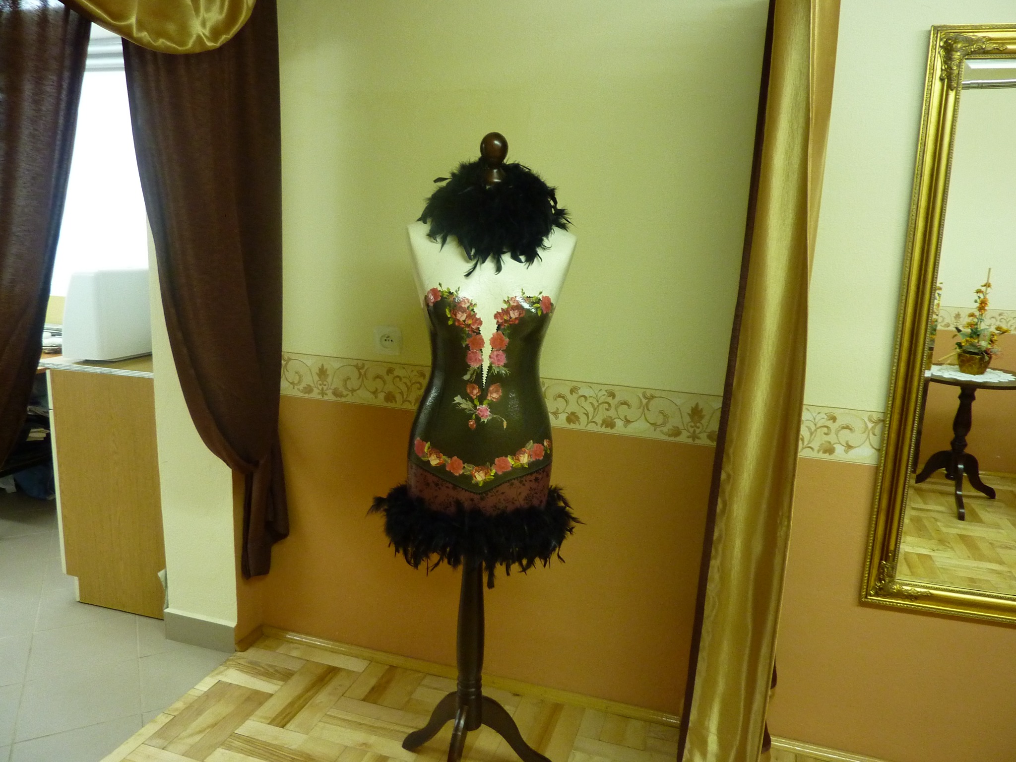 Manekin krawiecki z dekoracyjnym gorsetem w kwiaty i pióra, stojący w pomieszczeniu z beżowymi ścianami, drewnianą podłogą i złotą zasłoną