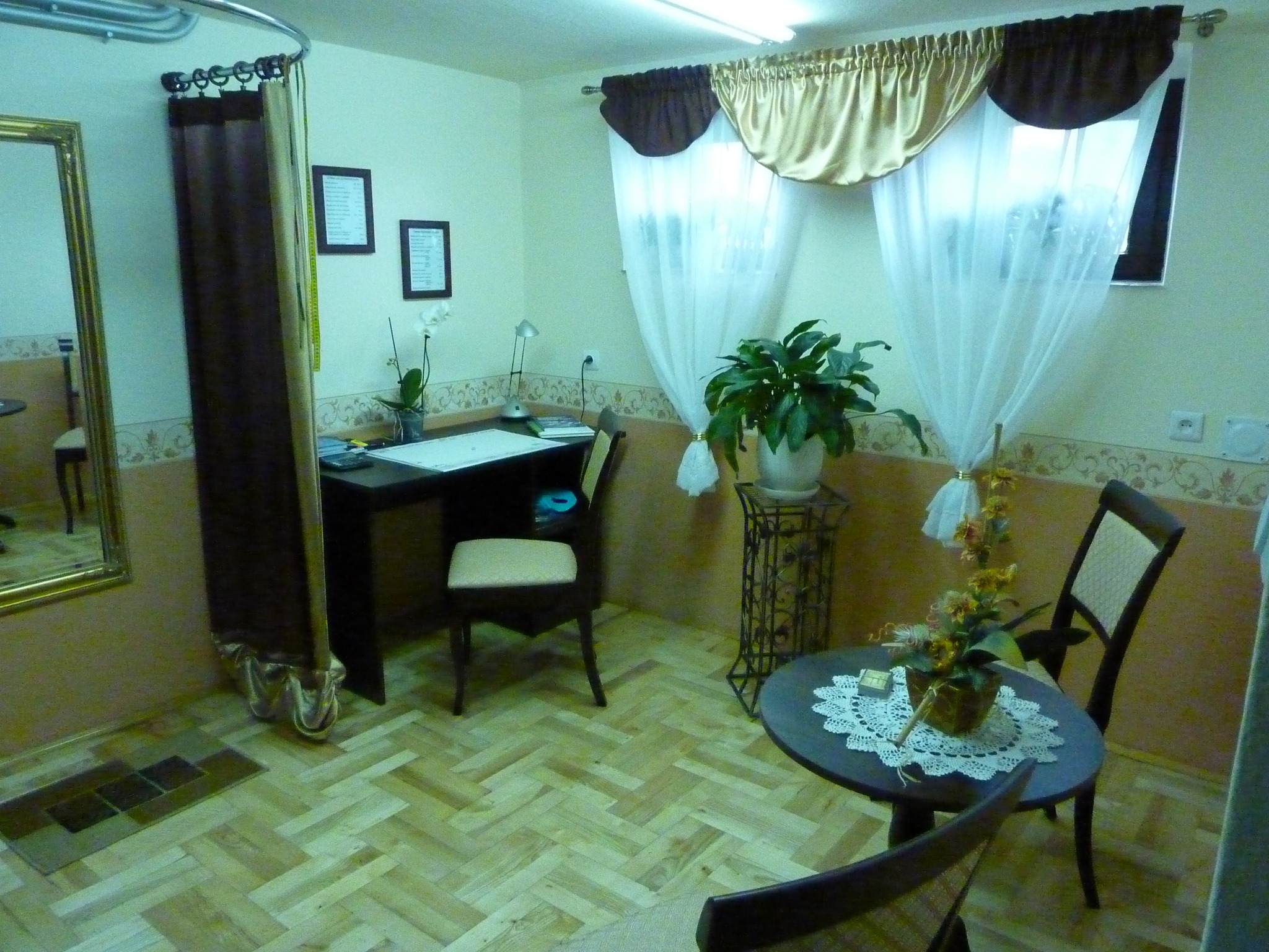 Wnętrze pracowni z biurkiem, krzesłem, lustrem i zasłonami, oświetlone sztucznym światłem, z roślinami doniczkowymi jako elementami dekoracyjnymi.