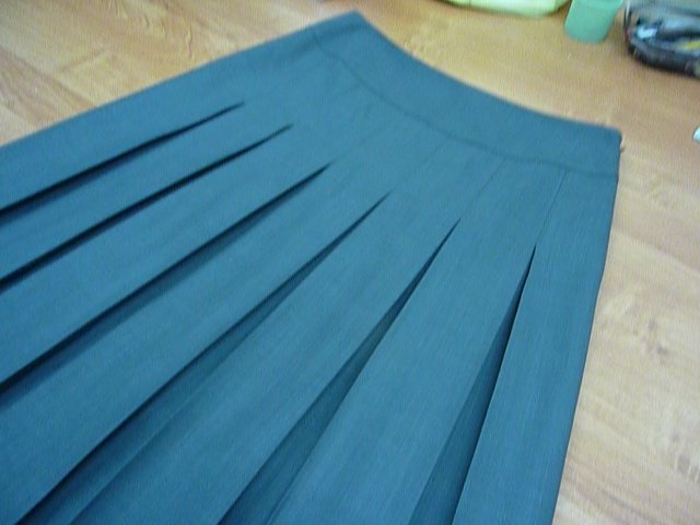 Fragment spódnicy z plisami w odcieniu ciemnego turkusu, leżącej na drewnianej podłodze, z widocznymi śladami szycia i wykończenia.