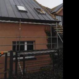 Dom jednorodzinny z częściowo wymienionym, ciemnym dachem z blachy trapezowej, widoczne dwa okna dachowe i rusztowanie przy ścianie budynku.