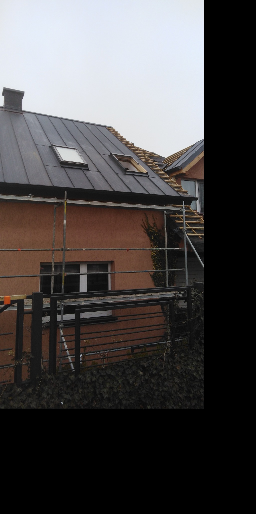 Dom jednorodzinny z częściowo wymienionym, ciemnym dachem z blachy trapezowej, widoczne dwa okna dachowe i rusztowanie przy ścianie budynku.