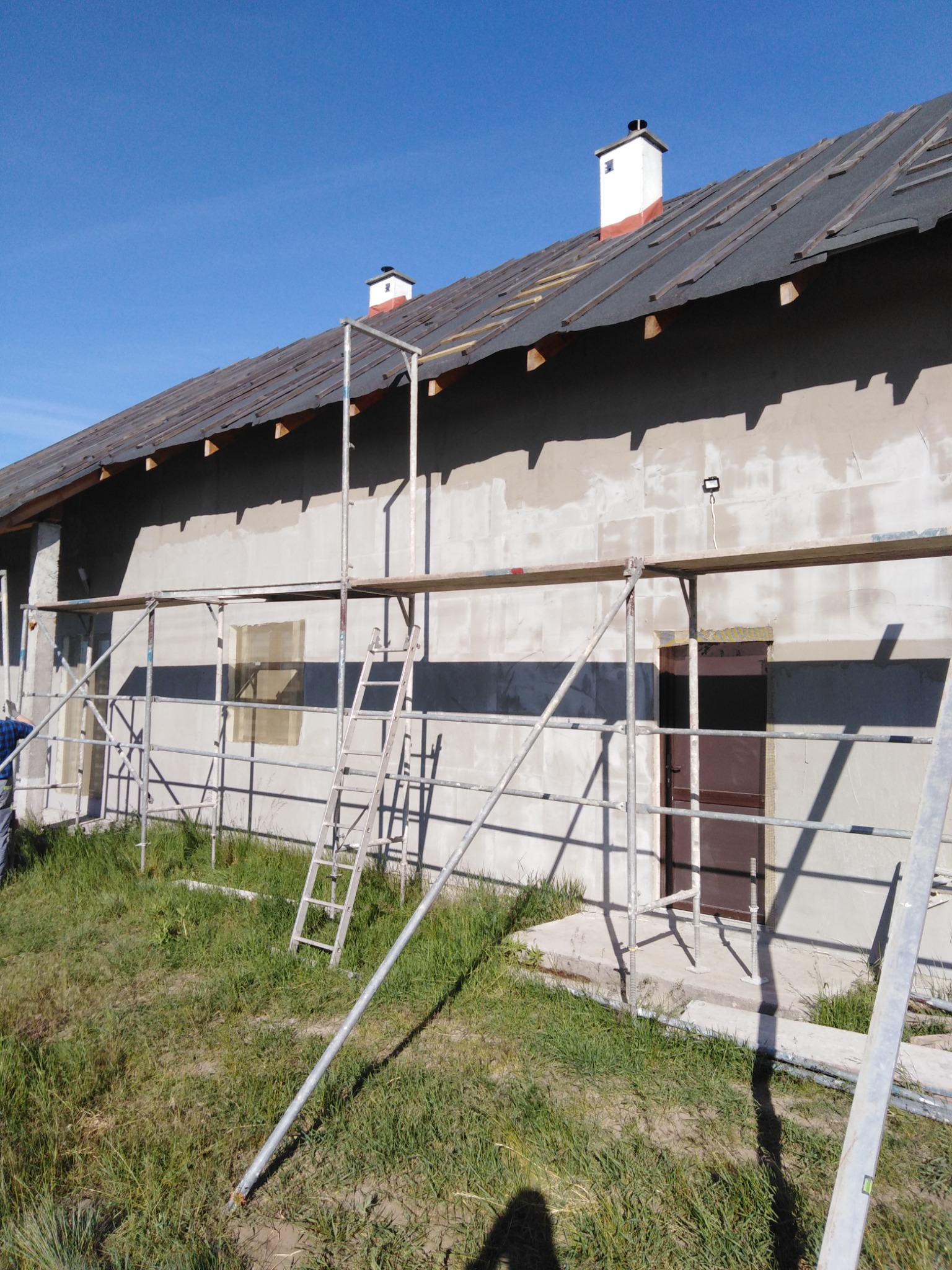 Dom w trakcie prac dekarskich, widoczne rusztowania, częściowo położona papa na dachu i kominy z białymi czapkami.