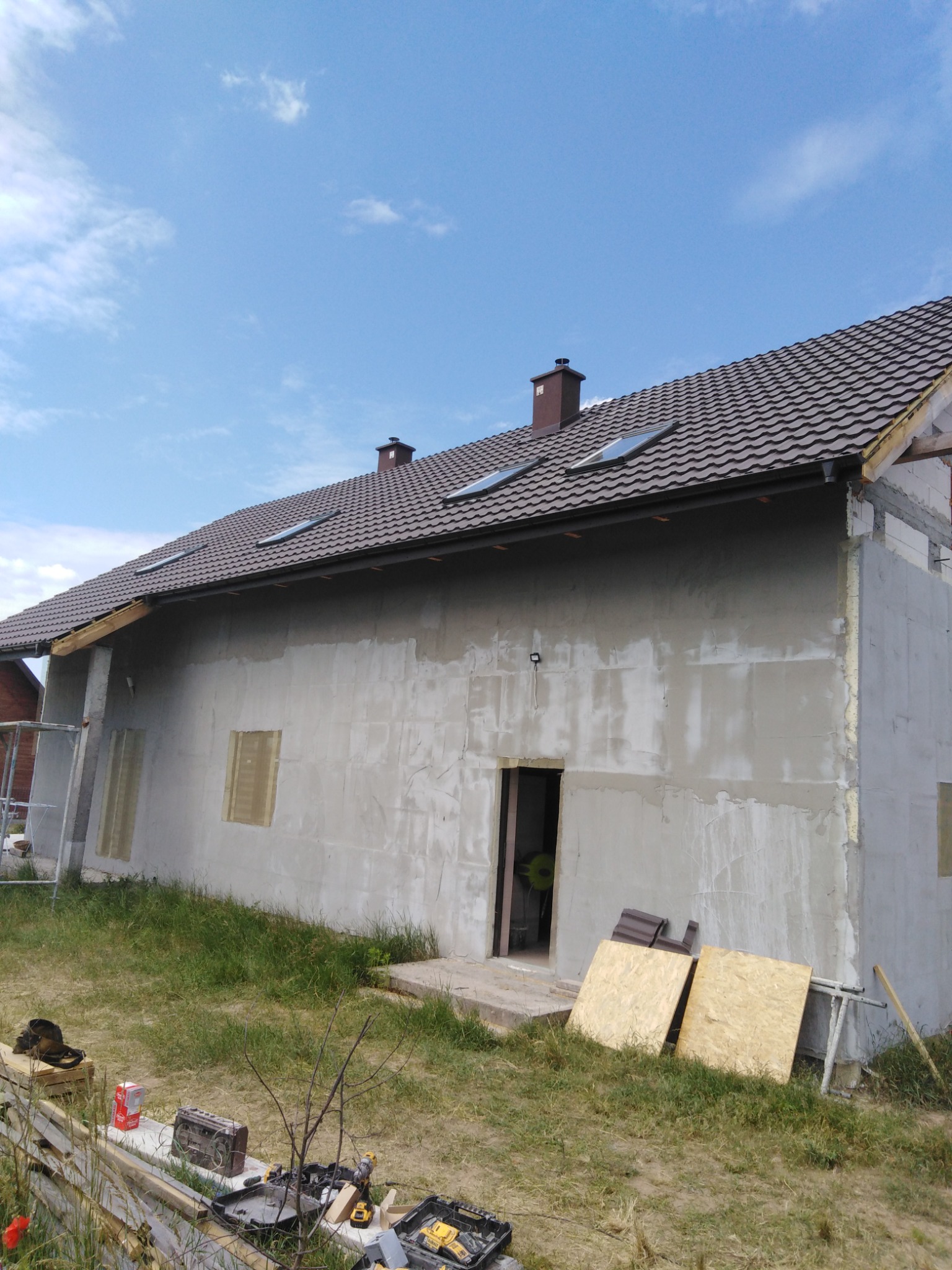 Dom w trakcie budowy z brązowym dachem z oknami dachowymi, surową elewacją i narzędziami budowlanymi na trawie.