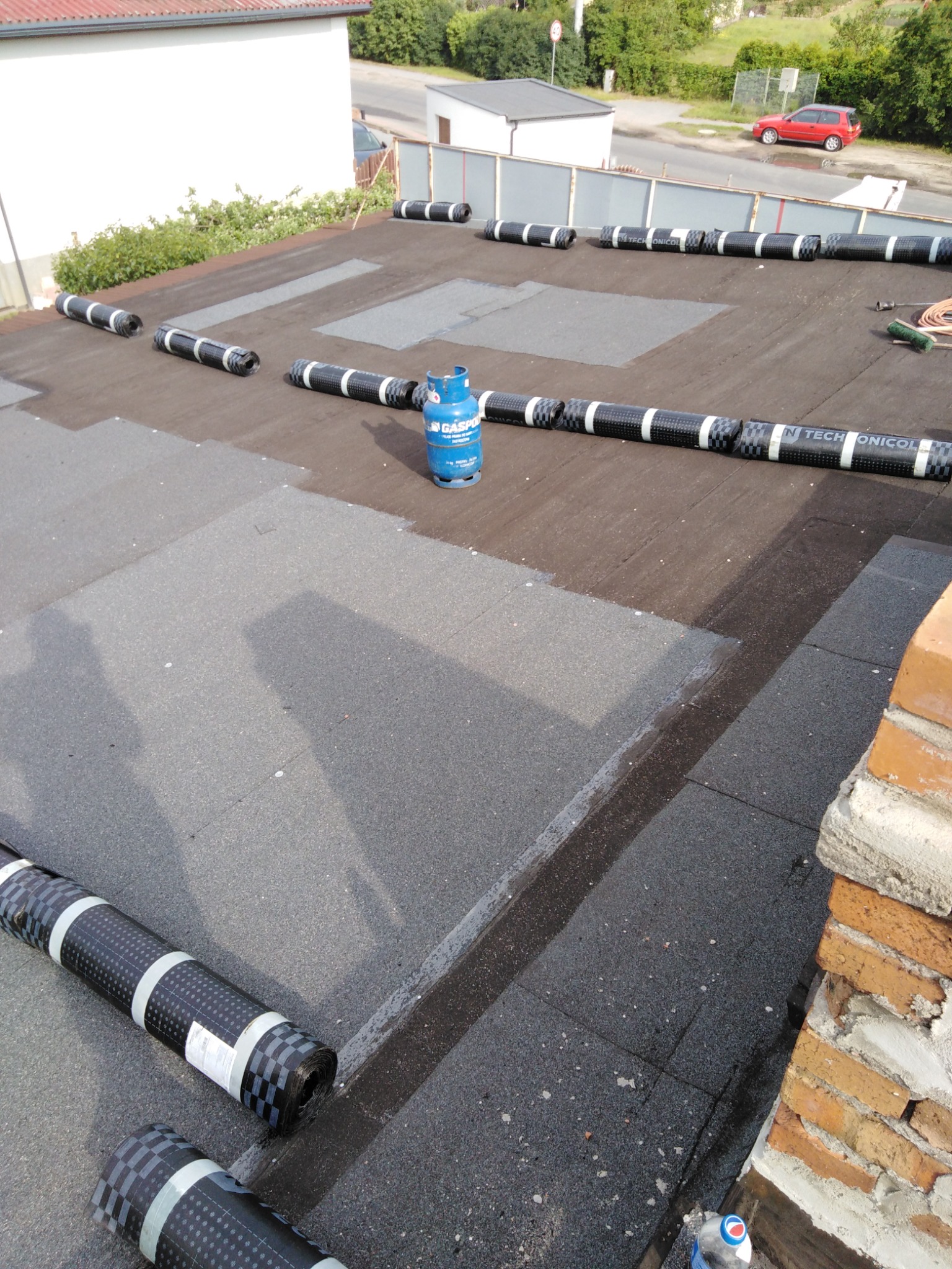 Widok z góry na płaski dach w trakcie prac: częściowo pokryty papą, z rolkami papy Techonicol i butlą gazową Gaspo na powierzchni, ceglana attyka w prawym dolnym rogu.
