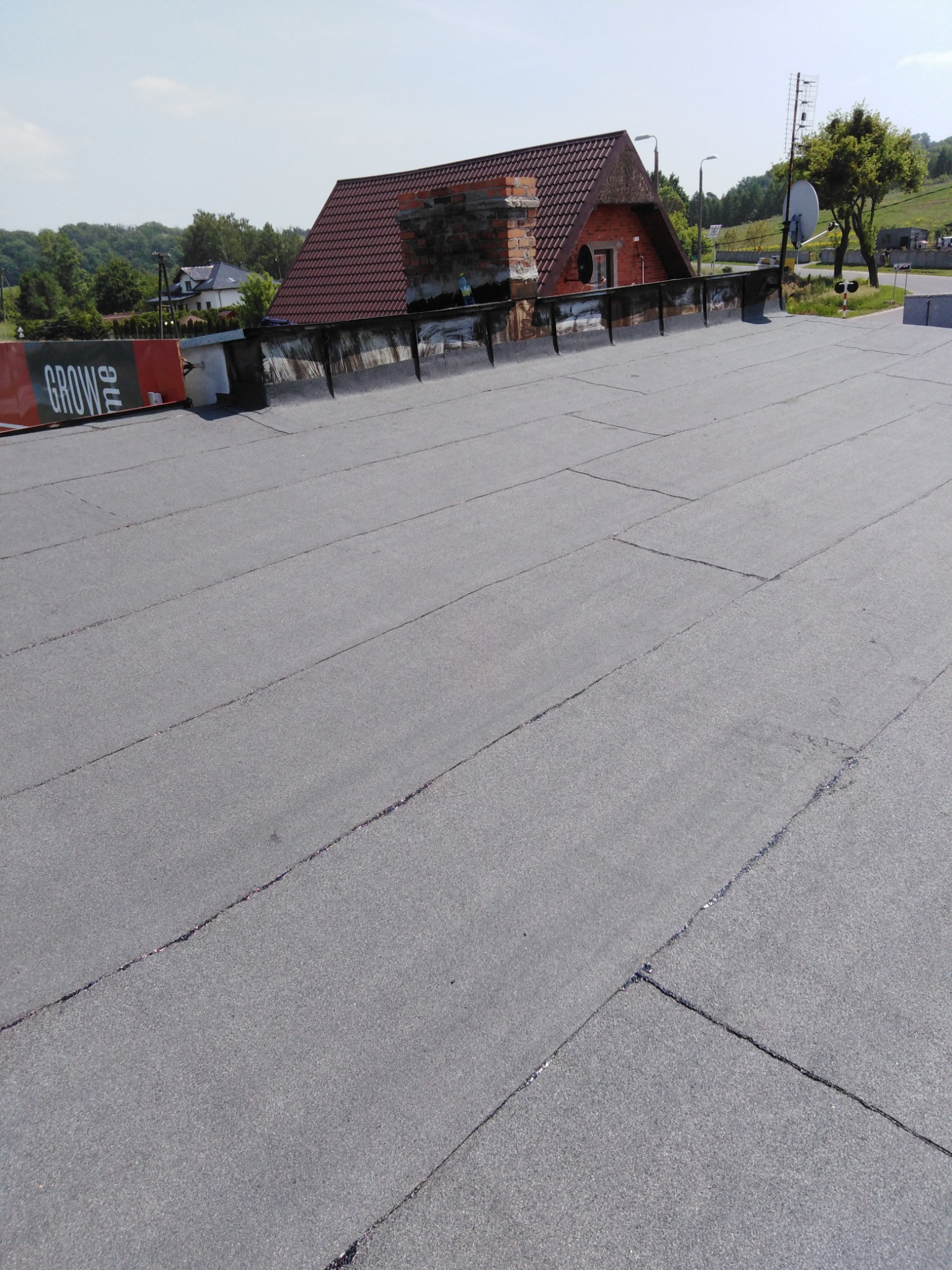 Szary, płaski dach pokryty papą termozgrzewalną z widocznymi łączeniami, na tle ceglanego domu z kominem i zielenią.