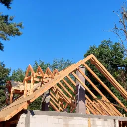 Wznoszenie drewnianej konstrukcji dachu na murowanym budynku w otoczeniu drzew, widoczny komin i pracownik na dachu.